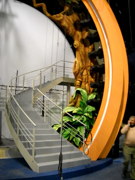 Detail of the tree trunk sculpted in the round flanking the portal: faux-stone bark with reliefs, large green tropical leaves rising from the base, white staircase with handrail climbing alongside