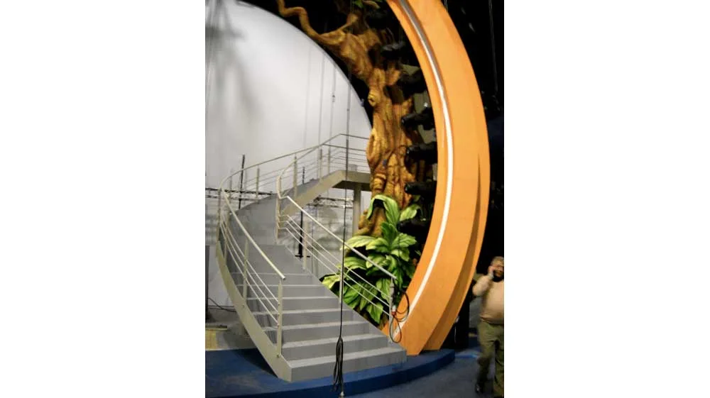 Side view of one of the Genodrome staircases: white polished metal spiral staircase climbing the flank of the portal, sculpted jungle tree trunk on the right with large tropical leaves, orange ring of the portal visible at the bottom right