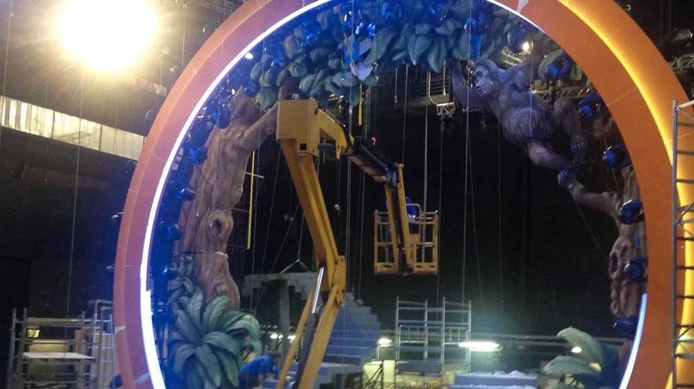 Night build of the portal with an industrial yellow cherry-picker at the centre lifting a worker for high-level finishes: sculpted jungle trunks on both sides, sculpted gorilla on the right climbing the trunk, orange portal ring, lighting from a single lamp top-left