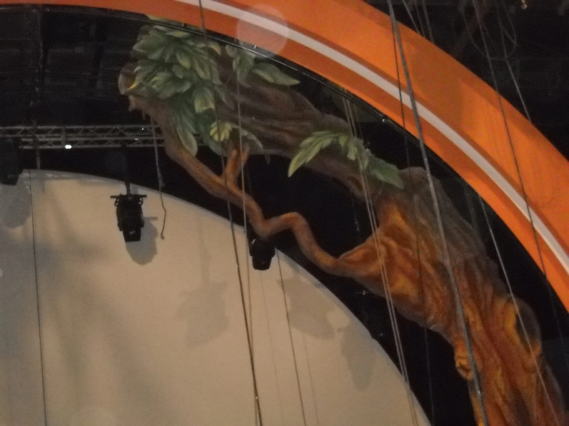 Ciao Darwin set during a night build: an industrial yellow cherry-picker at the centre lifting a worker for high-level finishes, view of the Genodrome portal on the right with sculpted trunk and tropical leaves