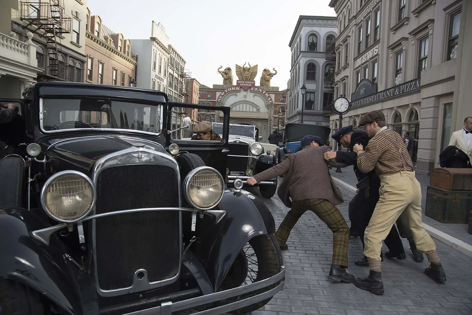 Cinecittà Street at the park: 1920s Manhattan façades, black Ford T cars parked along the street, actors in gangster and lady costume, Cabiria gate in the distance