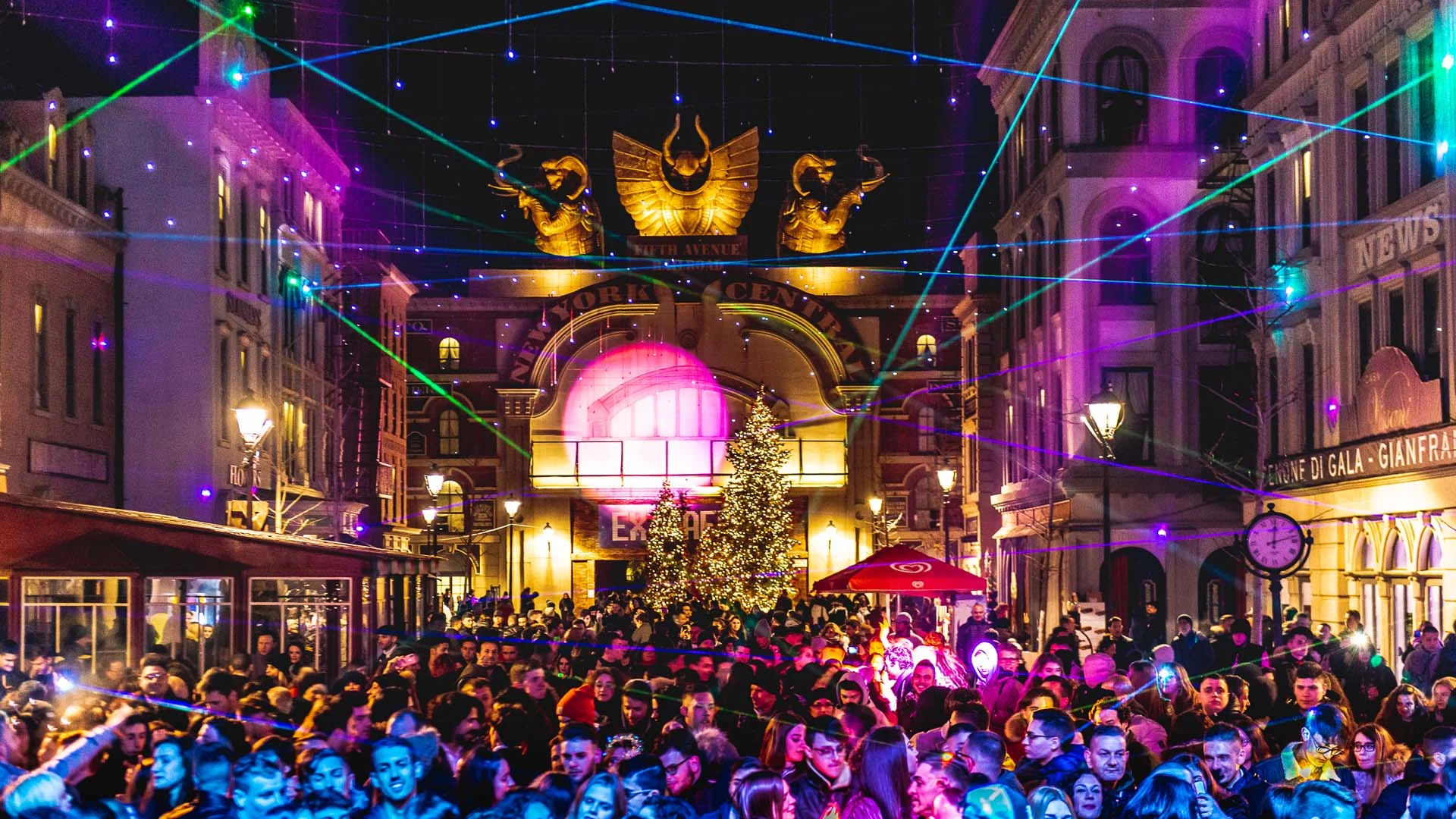 Cinecittà Street at night with laser show: illuminated arches hanging above the avenue, crowd of visitors, Christmas tree at the centre, lit-up façades