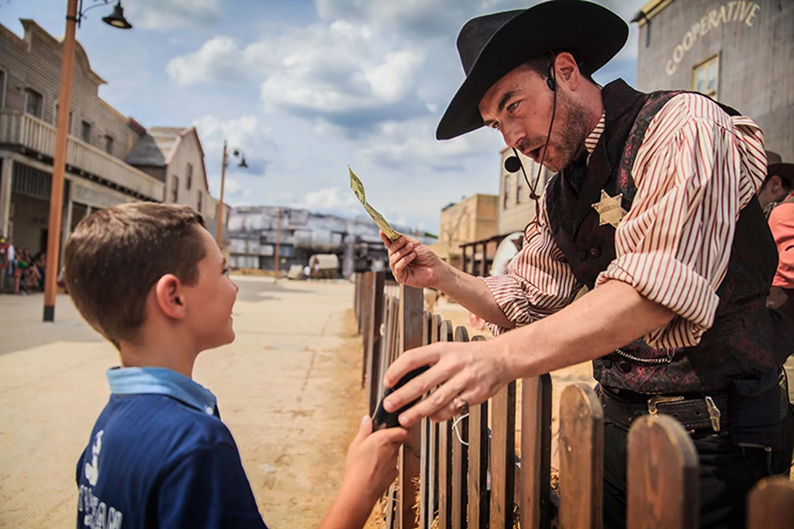 Actor in sheriff costume with star on chest and black hat hands a banknote to a child over a wooden fence, with the village cooperative in the background