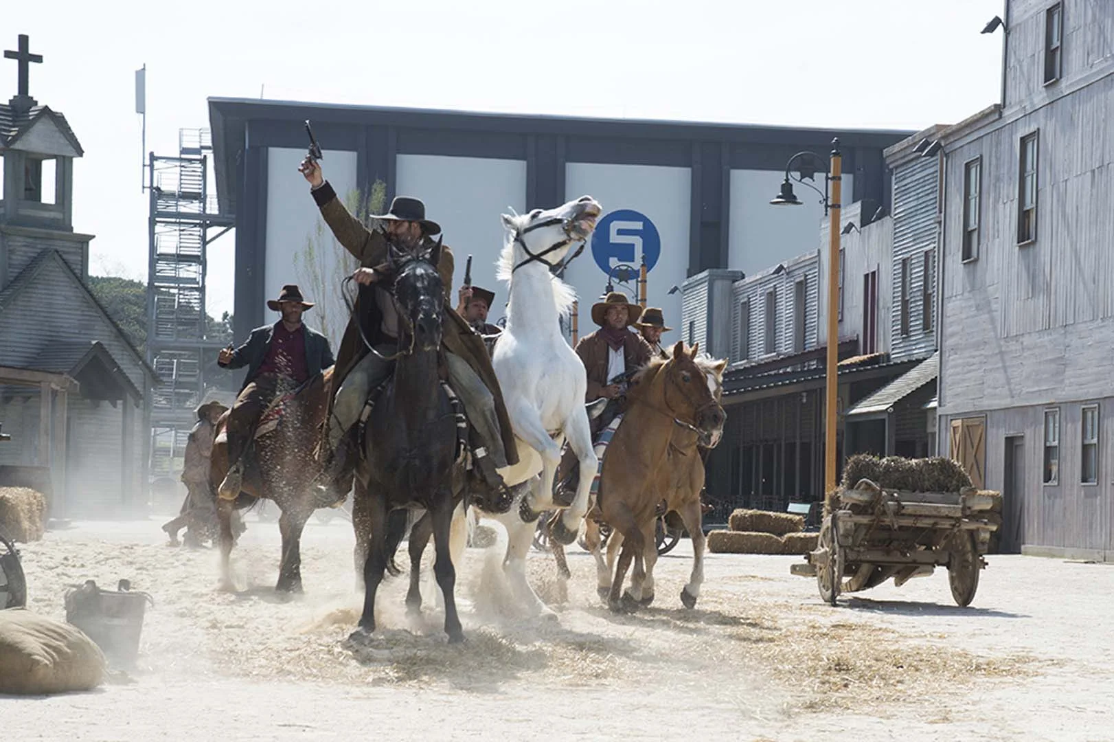 Far West set with five actors on horseback: gunslinger in the foreground firing with raised pistol, white horse rearing at the centre, two dark horses around, white dust, in the background a soundstage with a large '5' painted on the wall