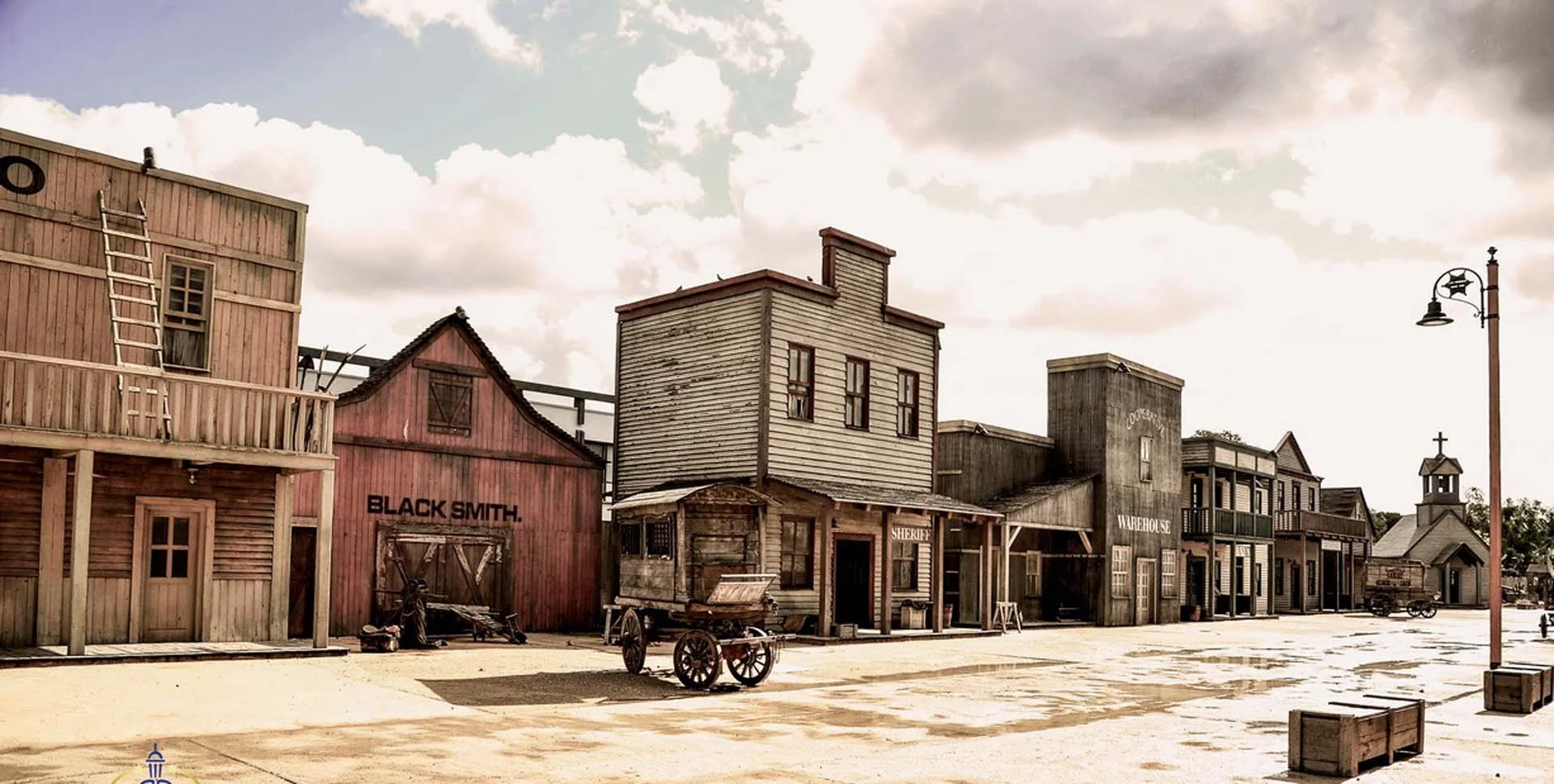 View of the Far West village at Cinecittà World: wooden buildings with 'Black Smith', 'Sheriff', 'Warehouse' signs, church with bell on the right, stockades, wooden cart at the centre, white dust on the ground