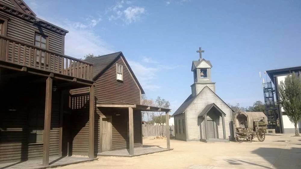 The Far West village at Cinecittà World photographed for the Creation Art portfolio: dark wooden buildings with overhang, first-floor balcony above a shop; white church with small bell tower and cross on the right; wooden cart between the buildings; metal frame of the haunted house visible at the back right