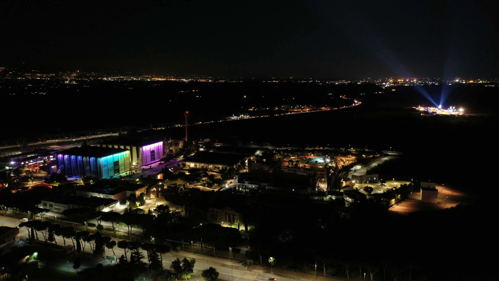 Aerial night view of Cinecittà World: Teatro 1 illuminated in rainbow colours, lit walkways, lights of Rome on the horizon