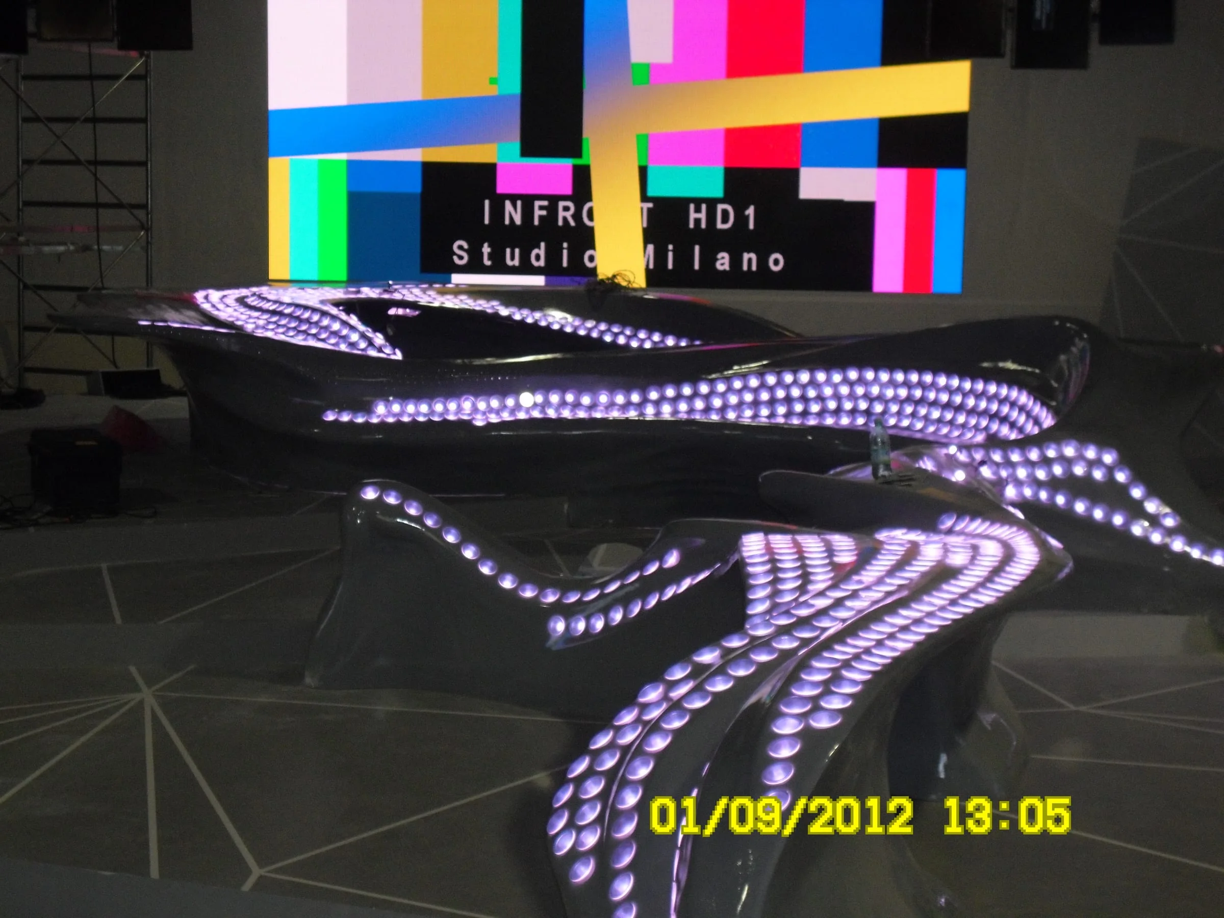 The Cristina Parodi Live desk on air at the Infront Studios in Milan: a sinuous gloss-black form with thousands of purple LEDs lit in concentric bands, HD signal test monitors in the background