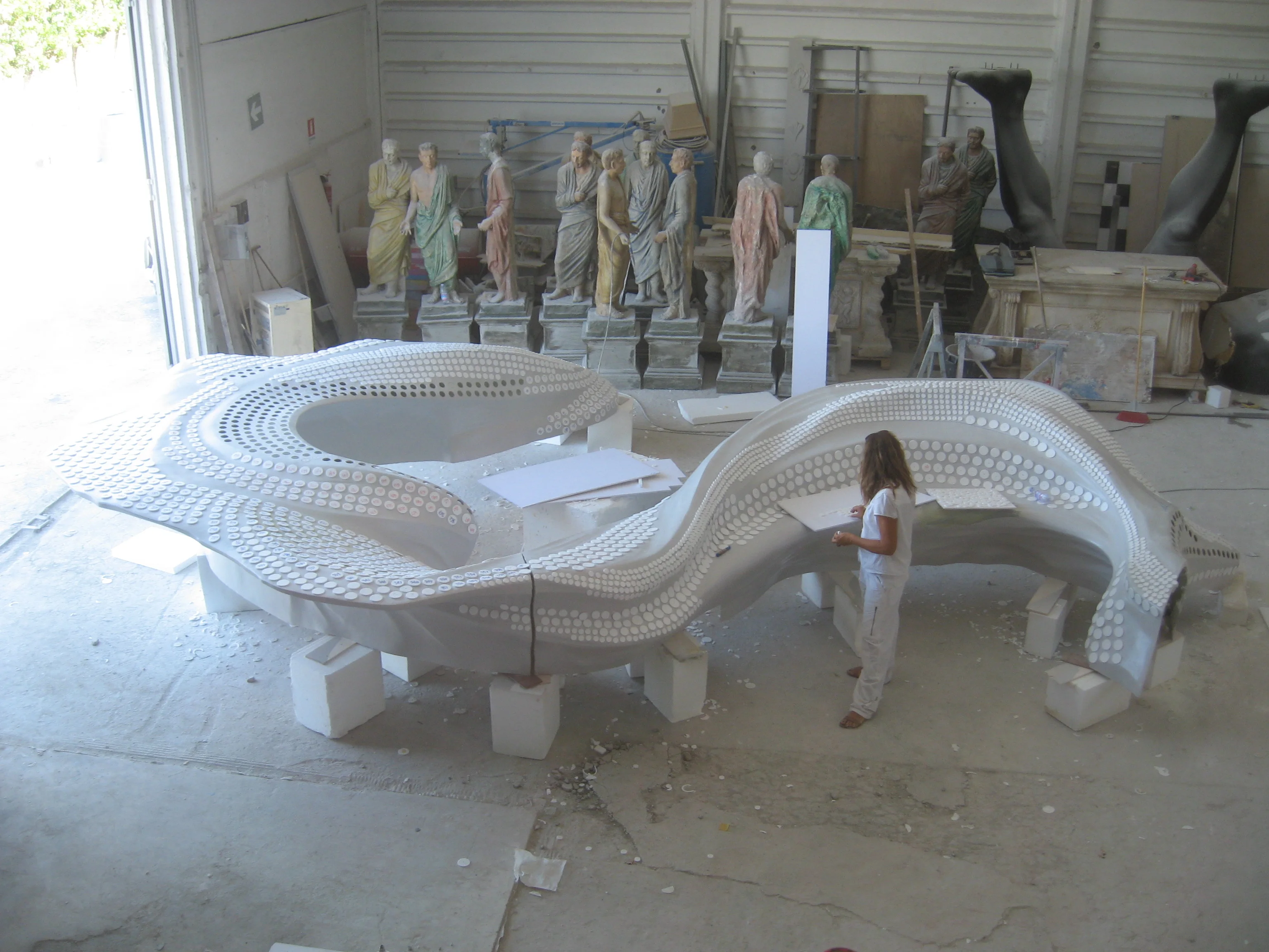 The desk being sculpted in our workshops in Marino: white polystyrene form worked by hand, the point-grid perforations already stamped on the surface, Roman statues from another set waiting in the background, a worker shaping the edge of the form