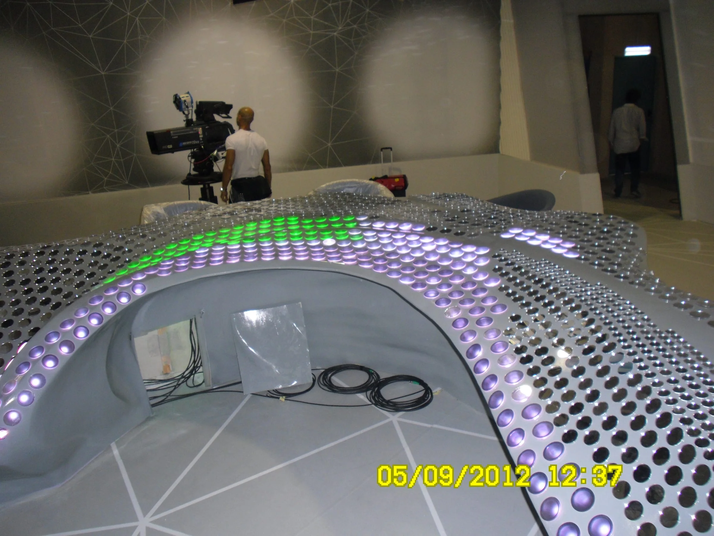 Detail of the desk in studio during LED programming: green and purple light points lit across the grey primer surface, camera operator and technician standing in the background of the Cristina Parodi Live set