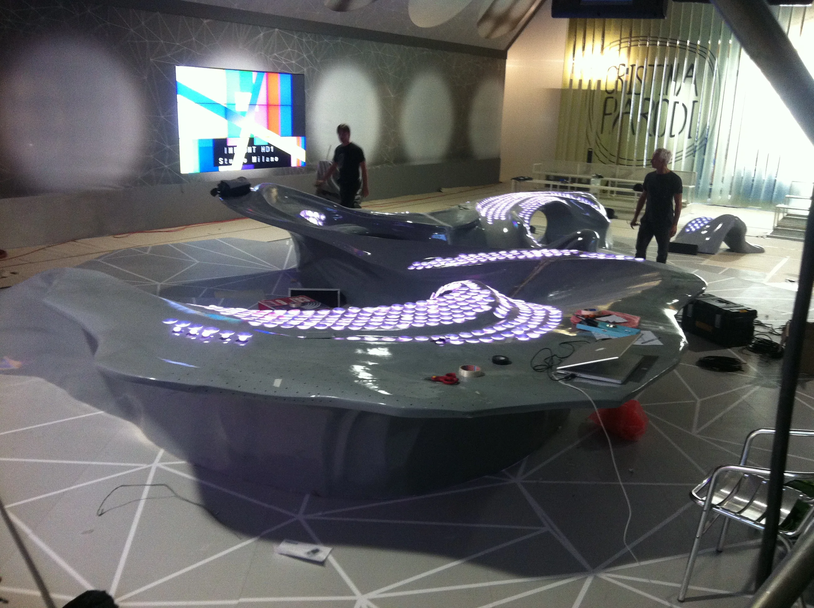 Panoramic view of the Cristina Parodi Live set at the Infront Studios in Milan: sinuous grey desk with purple LEDs lit at the center of the studio, technicians at work around it, PARODI lettering on the set backdrop