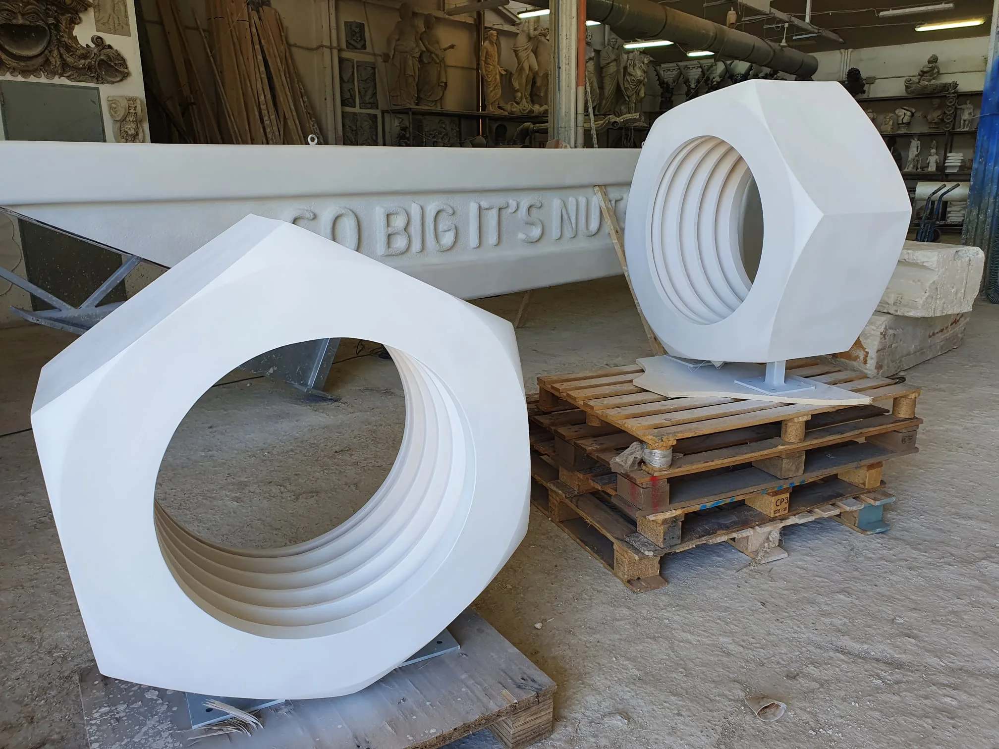 I due bulloni esagonali giganti scolpiti in polistirolo bianco, vista ravvicinata: forma esagonale con foro centrale per la chiave inglese, scolpiti a tutto tondo, posti su pallet di legno nel piazzale del laboratorio, accanto la parte iniziale della chiave inglese con la scritta in bassorilievo