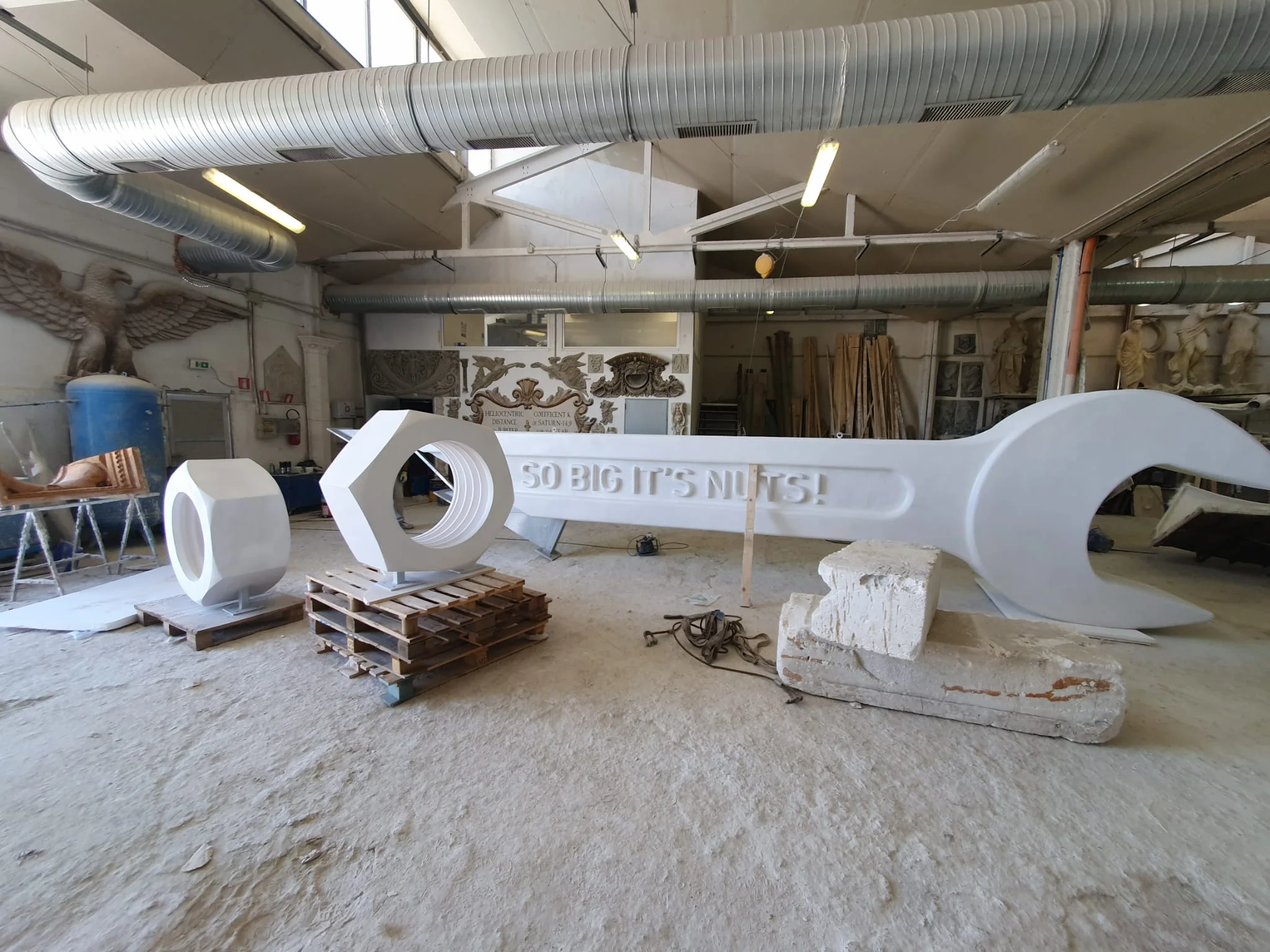 Chiave inglese gigante scolpita in polistirolo bianco nel laboratorio Creation Art, lunga circa quattro metri, posata orizzontalmente con la scritta «SO BIG IT'S NUTS!» in bassorilievo sul manico, accanto due bulloni esagonali giganti uno appoggiato a terra e uno su pallet di legno; sullo sfondo statue classiche e fregi del nostro archivio