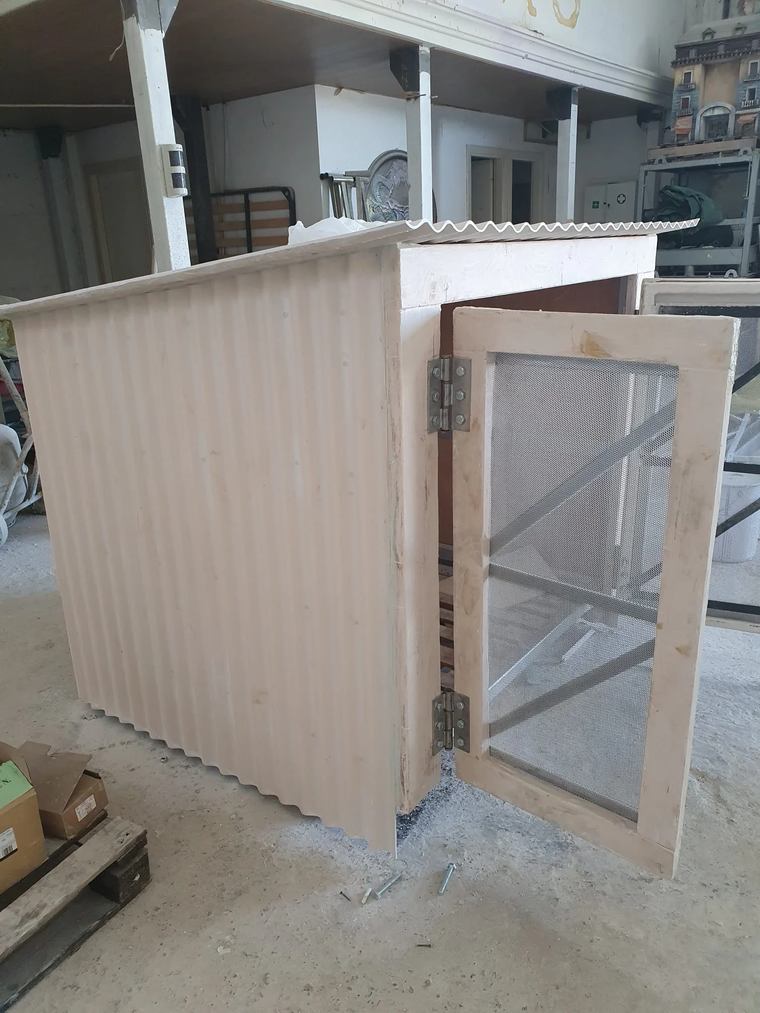 La casetta-officina con porte aperte mostrando l'interno scuro vuoto: struttura interna metallica visibile, pallet di legno sul pavimento, scaletta a pioli a sinistra, vista dall'esterno della facciata fronte