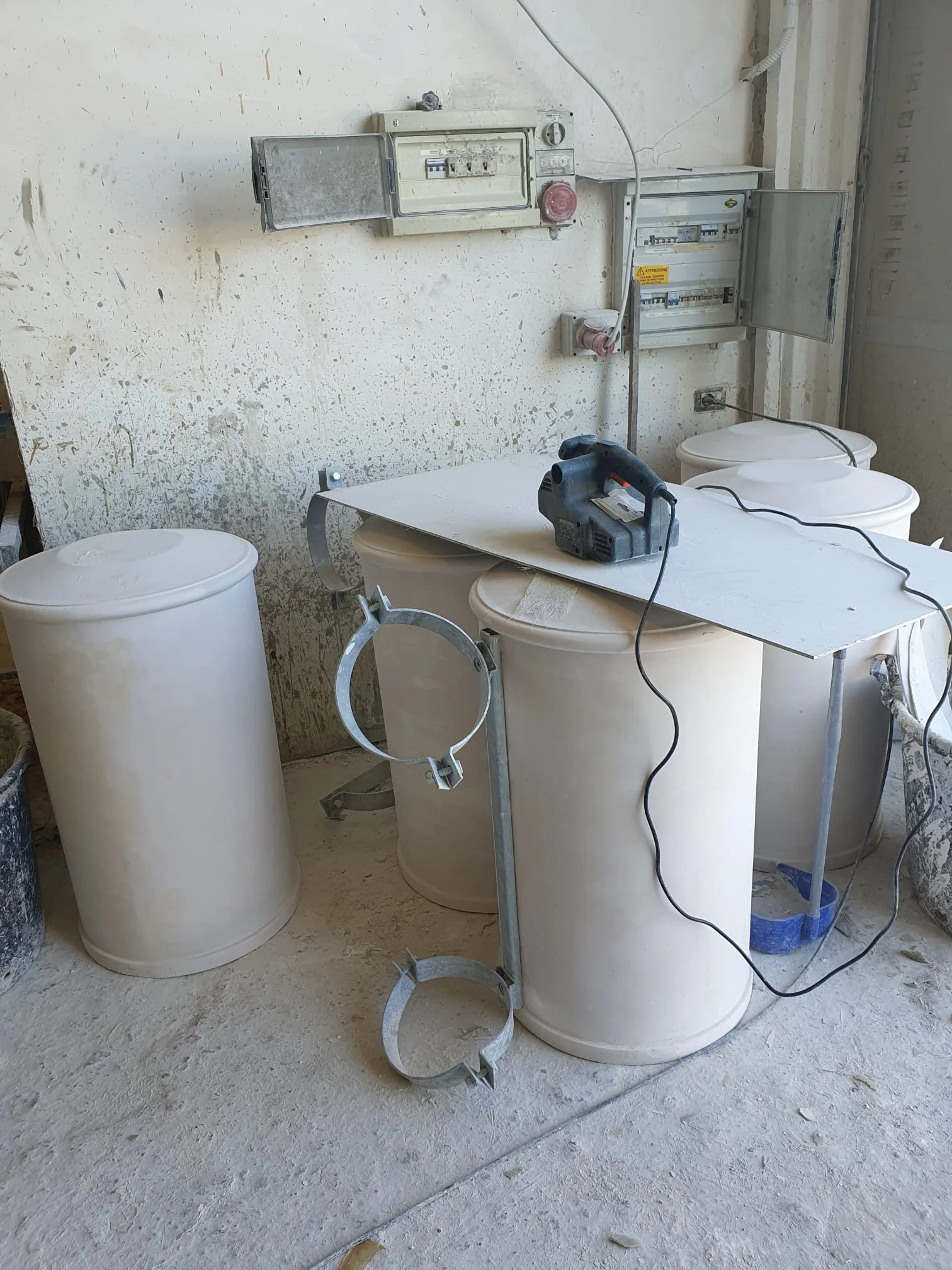 Casetta-officina scenografica in polistirolo, ancora nel laboratorio: tetto in lamiera ondulata grigia stilizzata, doppia porta in legno con rete metallica chiusa, pareti laterali a doghe corrugate, dimensioni circa un'altezza d'uomo e mezza, posata su pavimento del capannone con scala alle spalle
