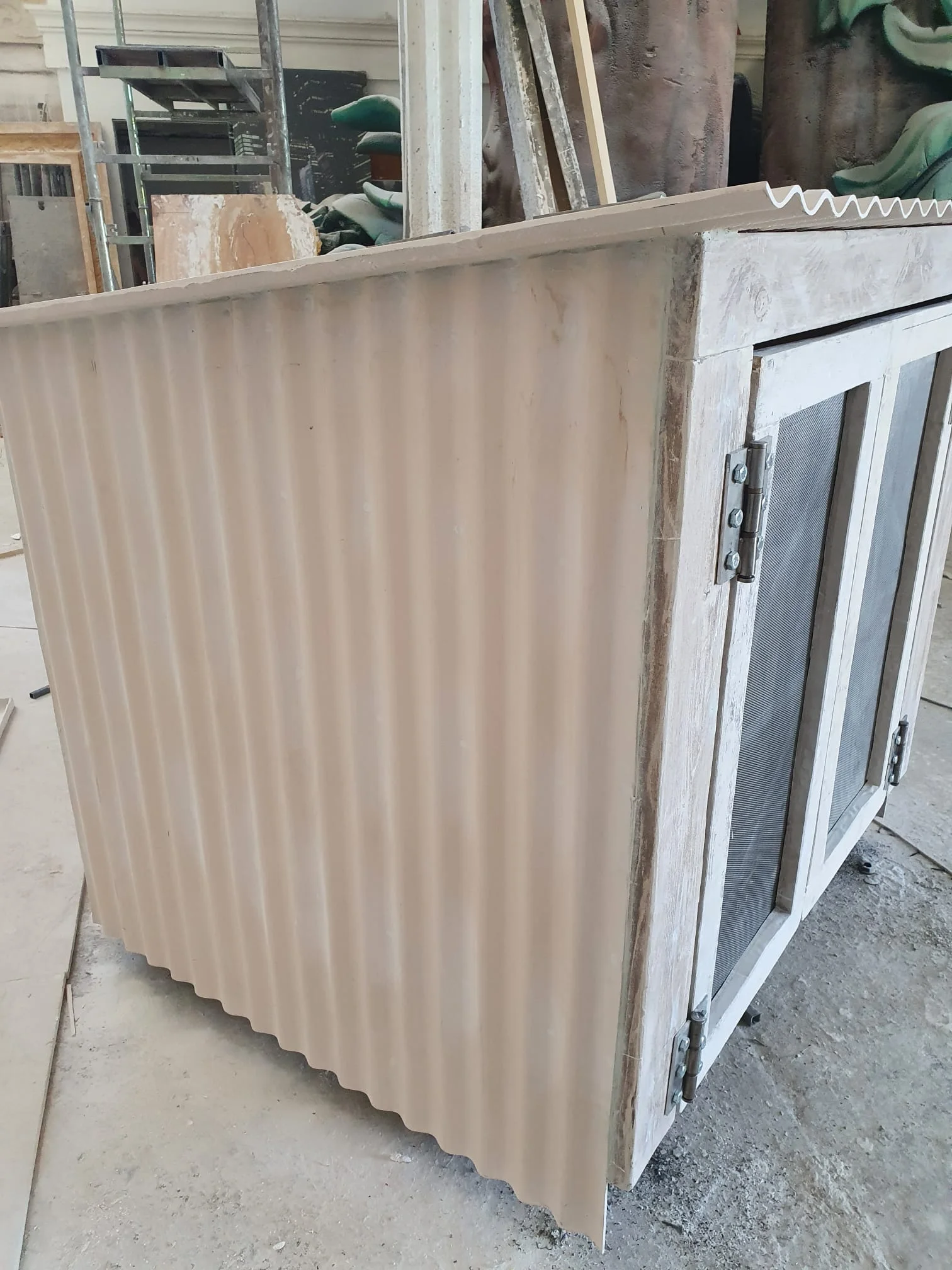 Vista dall'interno della casetta: pareti grigio scuro con struttura interna a graticcio metallico, pallet di legno sul fondo, pavimento del laboratorio in cemento polveroso, struttura aperta come una vetrina di esposizione
