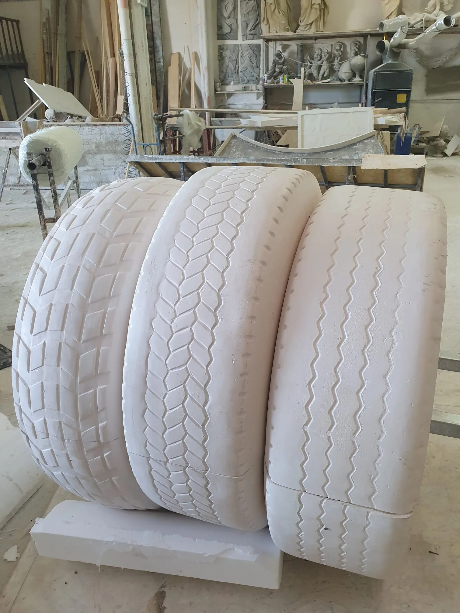Tre pneumatici giganti scolpiti in polistirolo bianco grezzo, ognuno con disegno di battistrada diverso (uno a tasselli quadrati, uno a tasselli esagonali, uno a strisce orizzontali), posati di taglio nel laboratorio sui pallet di legno con sullo sfondo statue classiche del nostro magazzino