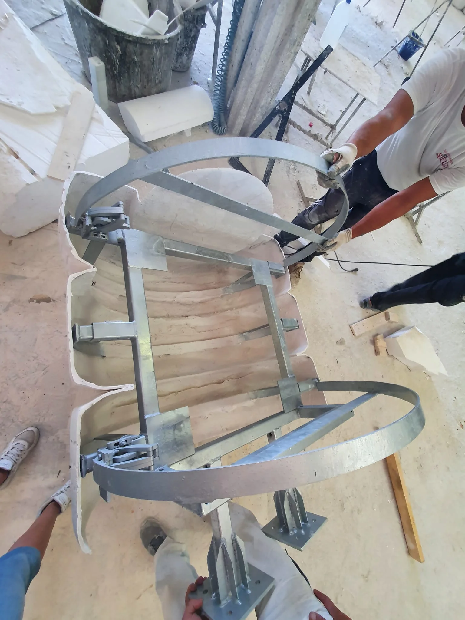 Dettaglio del montaggio dei cerchi metallici di rinforzo del silos: due operai in tuta da lavoro stanno assemblando le due bande circolari in acciaio che racchiudono il guscio del silos, vista dall'alto, frammenti di polistirolo sparsi sul pavimento