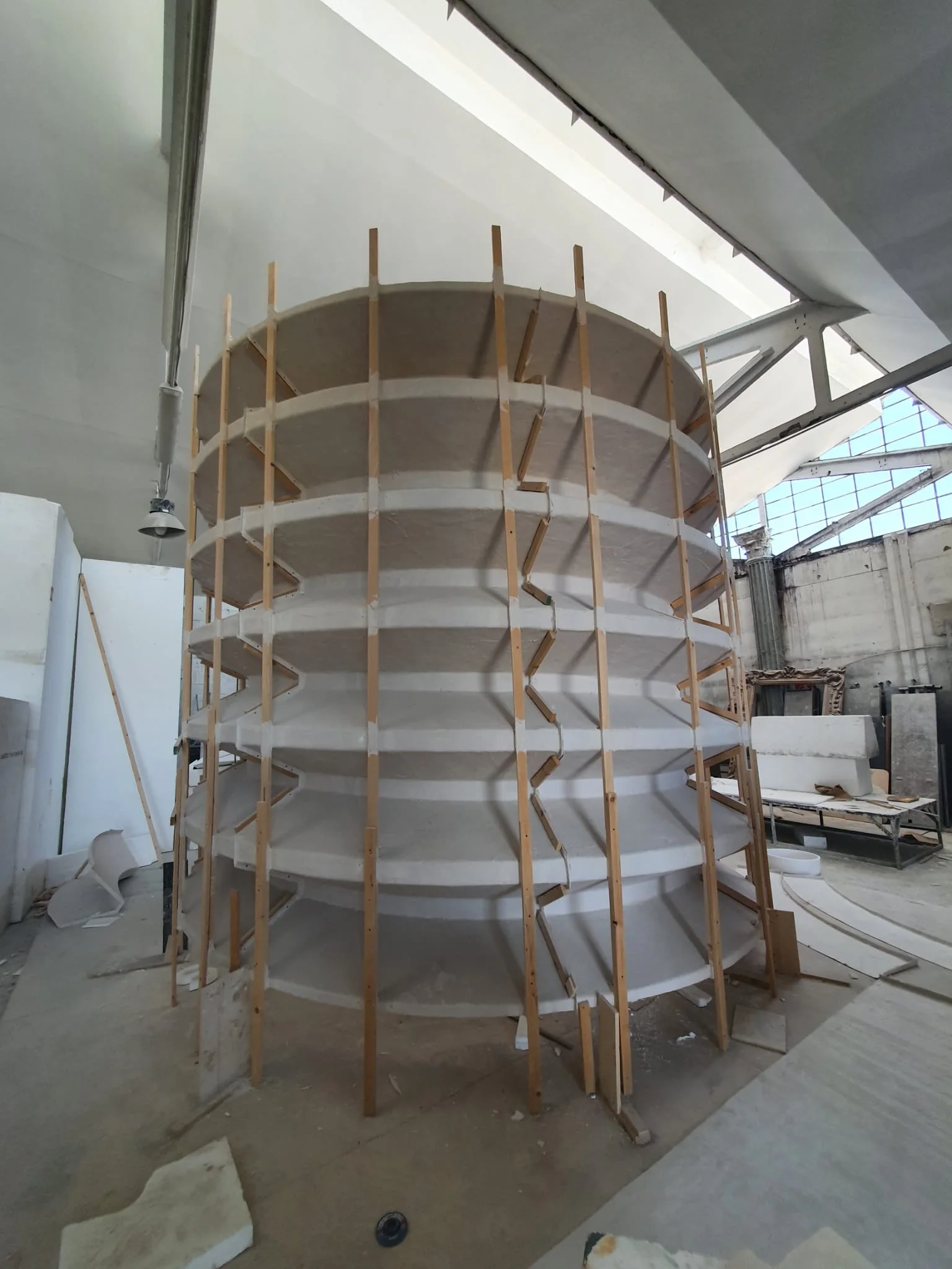 Grande silos cilindrico stratificato a piani sovrapposti scolpito in polistirolo bianco grezzo, alto circa tre metri, struttura cilindrica con cerchiature orizzontali a costole e listoni verticali in legno di rinforzo, posato nel laboratorio con vetrate in lontananza