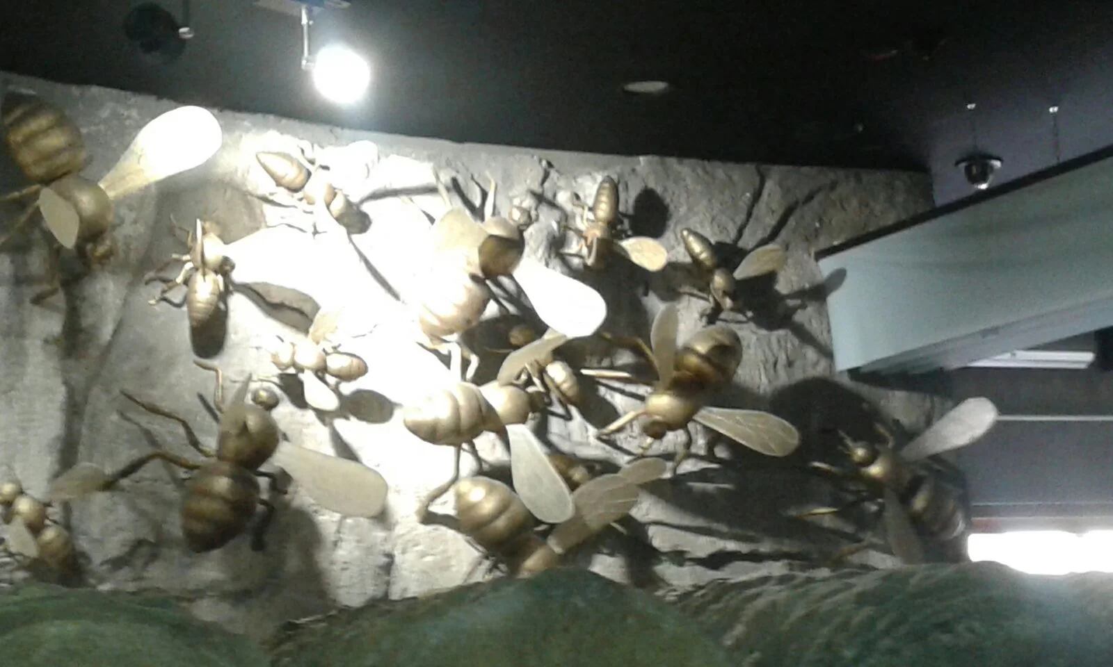 Scenographic panel with ten giant golden bees sculpted in the round on a grey rock wall for Expo Milano 2015: each bee about a metre long, golden banded body, translucent white wings, articulated legs, arranged against a faux-stone wall backdrop with reliefs of dry branches