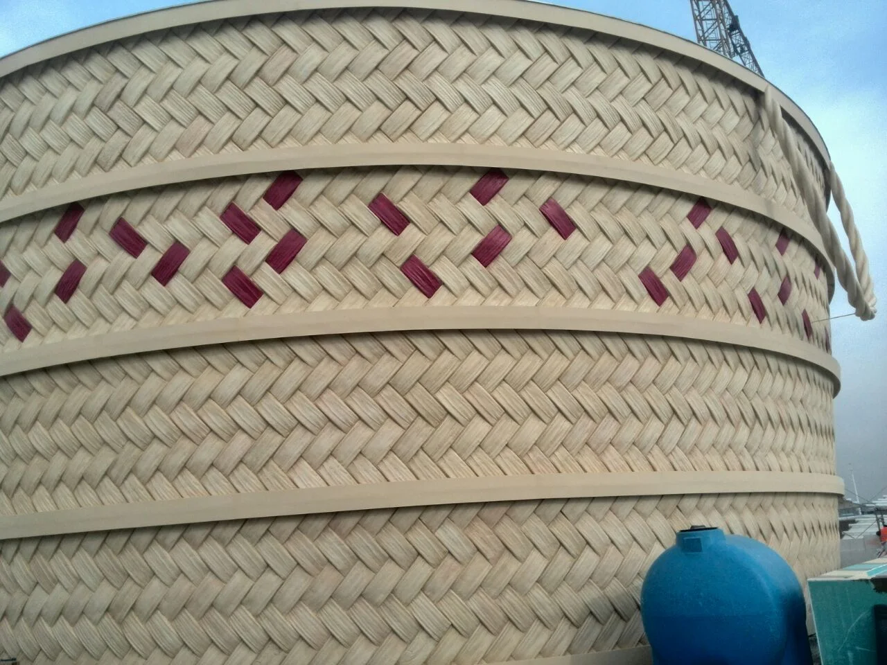 Detail of the woven facade of the Qatar Pavilion: the fibreglass-recreated surface reproduces the pattern of the traditional Jereer, five shades of straw-coloured thread with horizontal bands and a central band of burgundy diamonds from the flag; a yellow construction crane visible at top right, a blue tank at bottom right