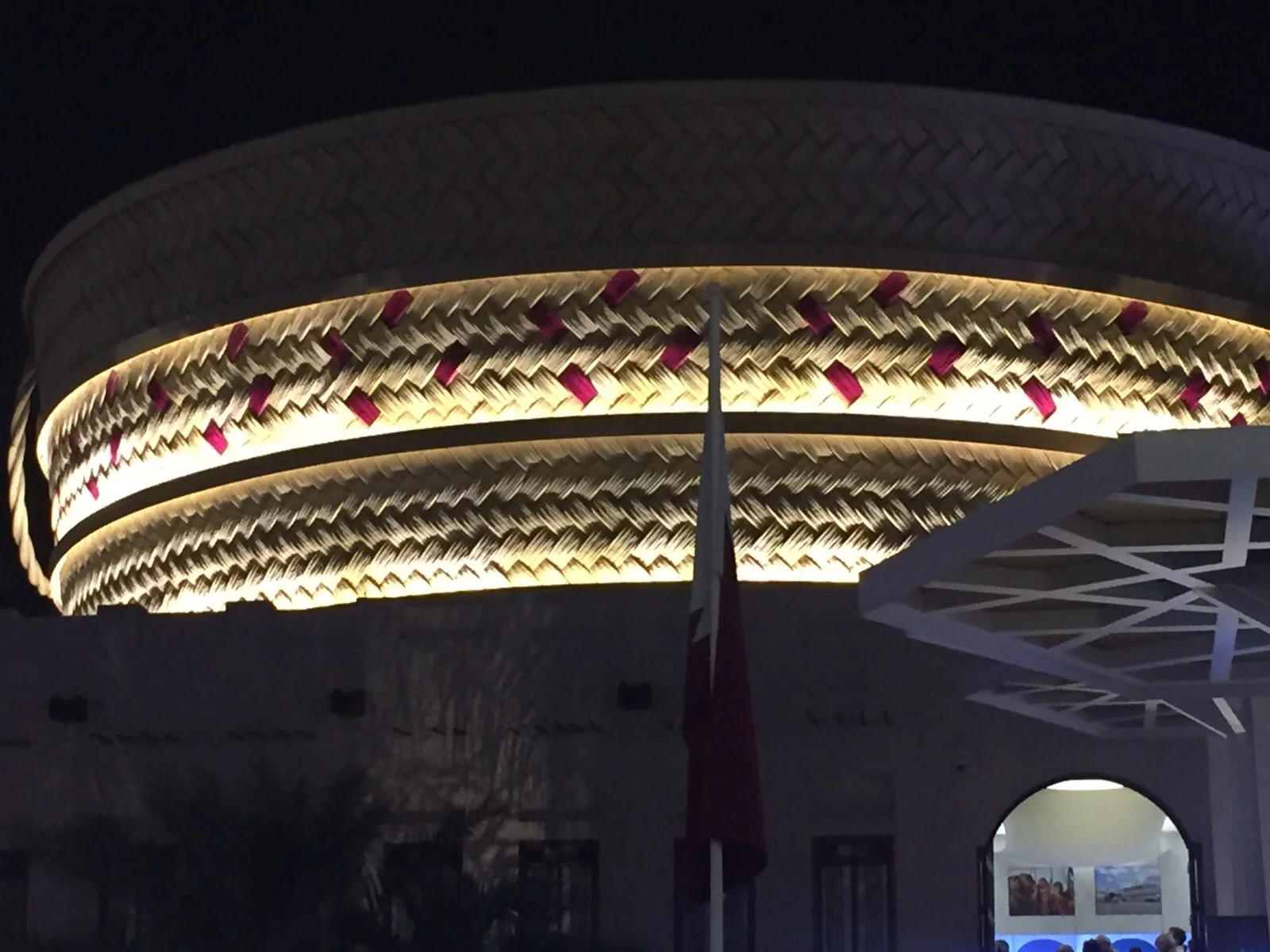 Qatar Pavilion at Expo Milano 2015 seen at night: the curved facade is covered by the Jereer cladding — woven texture in straw colour with burgundy inserts of the Qatari flag — lit from within; in the foreground the Qatar flag flying from a flagpole, the white arched entrance to the right
