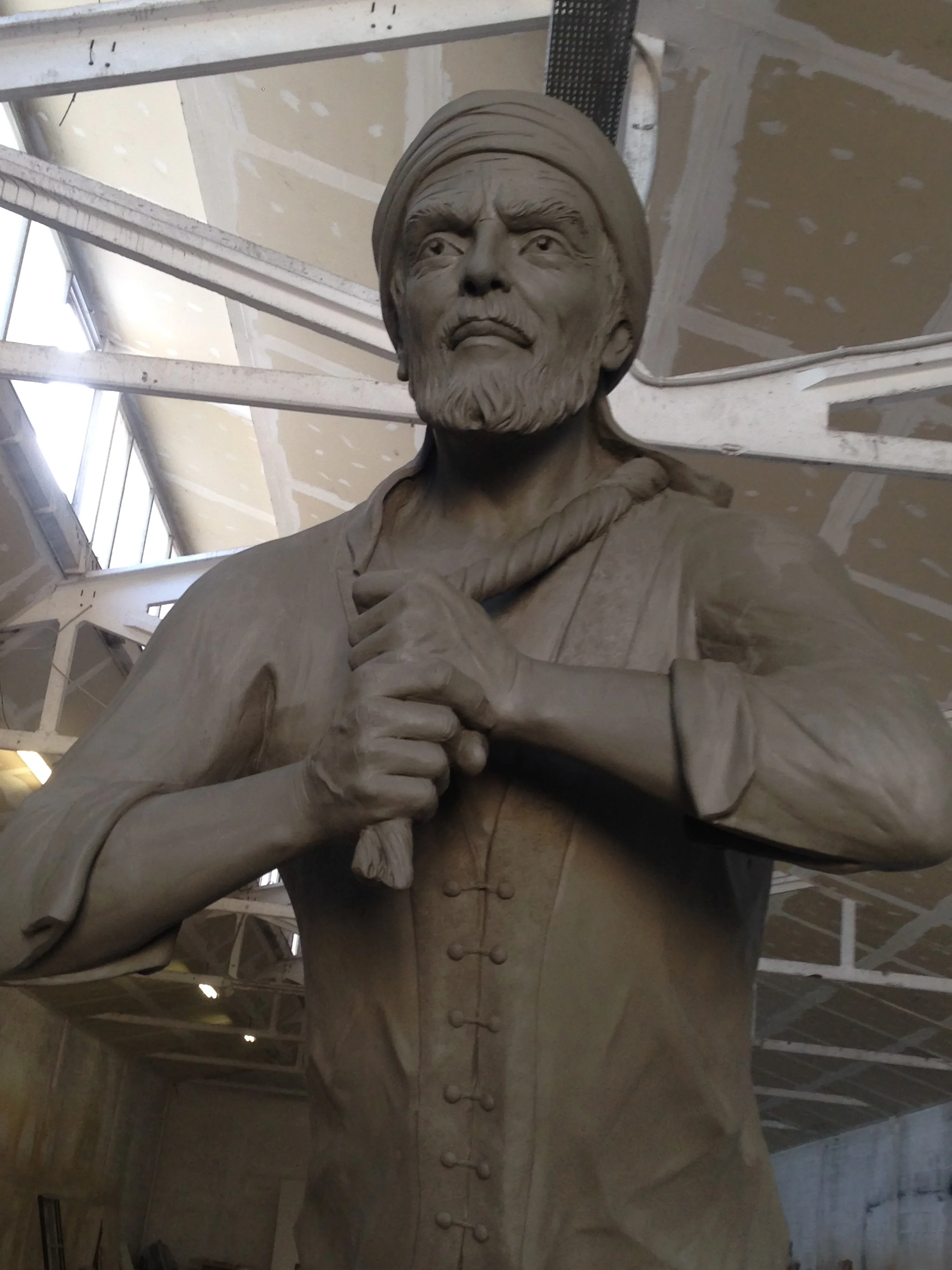 The statue in front view, legs slightly apart in a walking pose, right hand at the chest, keffiyeh modelled, bearded face turned to the right; workshop walls in the background with other partial sculptures