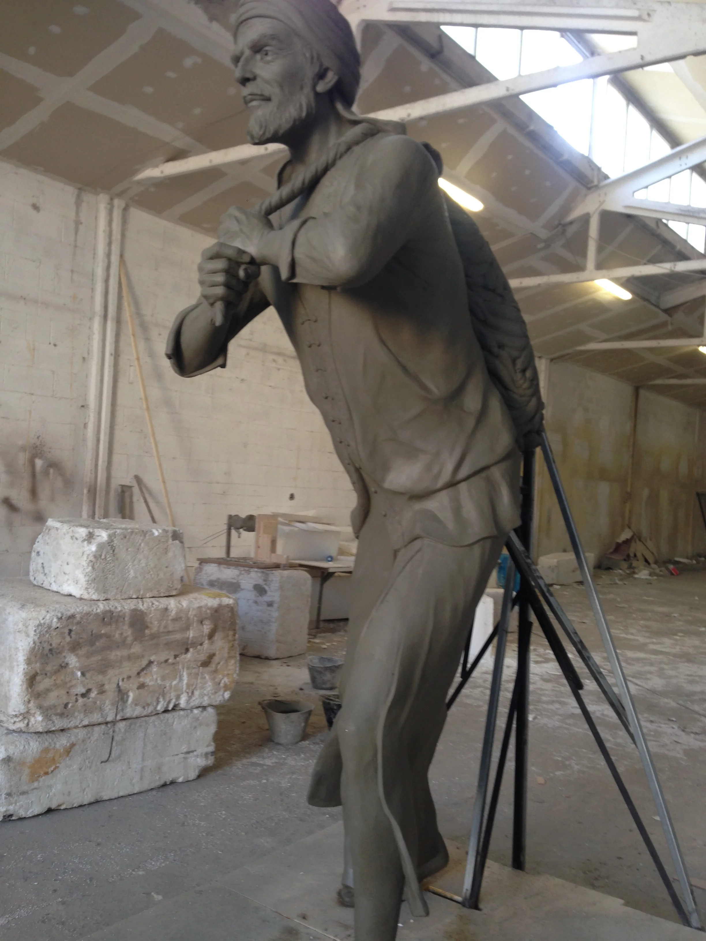 Three-quarter view of the statue in the workshop: pose with the right arm raised to the chest, keffiyeh modelled over the head, long tunic falling to the feet, raw concrete base, workshop wall in the background