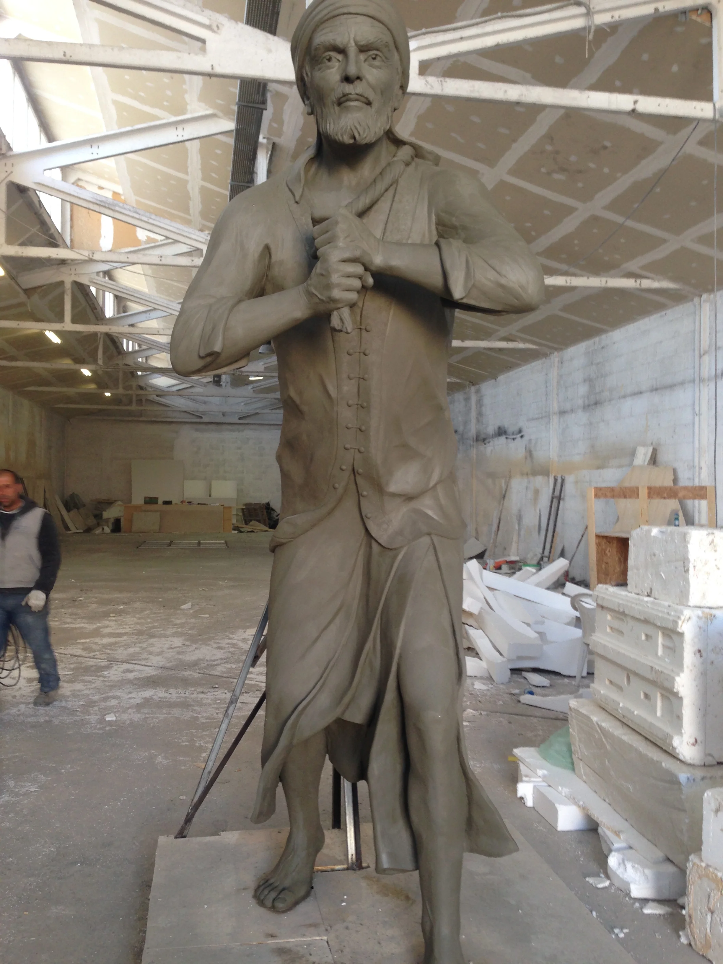 The statue at a more advanced stage of work, still sienna coloured: face modelled with the beard, shirt drapery refined with the spatula, hand anatomy defined; workshop hall with materials in the background
