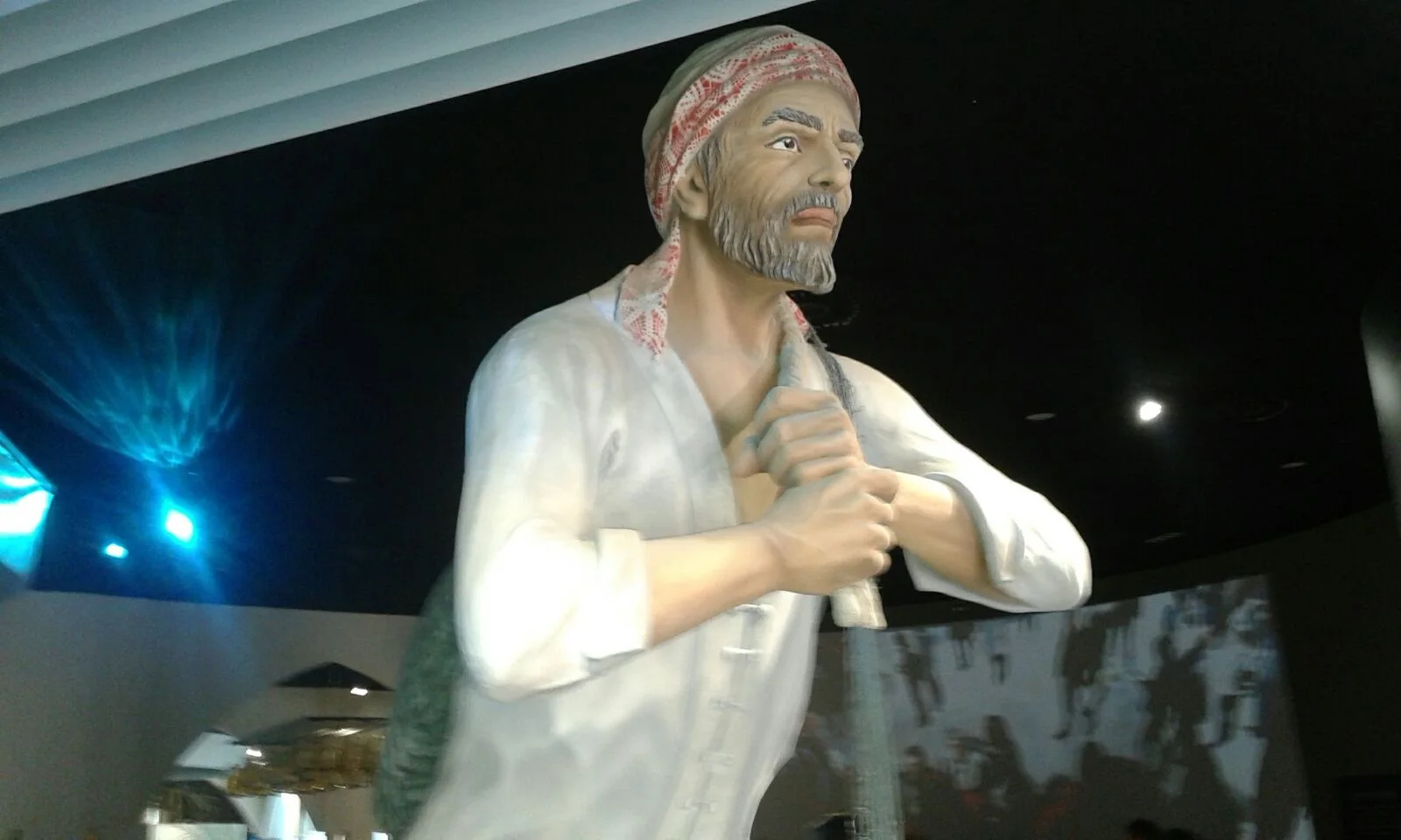 The statue of the Qatari man installed in the pavilion: fisherman-farmer with a white and red checked keffiyeh, buttoned white shirt, grey beard, blue eyes, proud expression, hands joined on the chest, behind an animated backdrop of images of the country; blue stage lighting