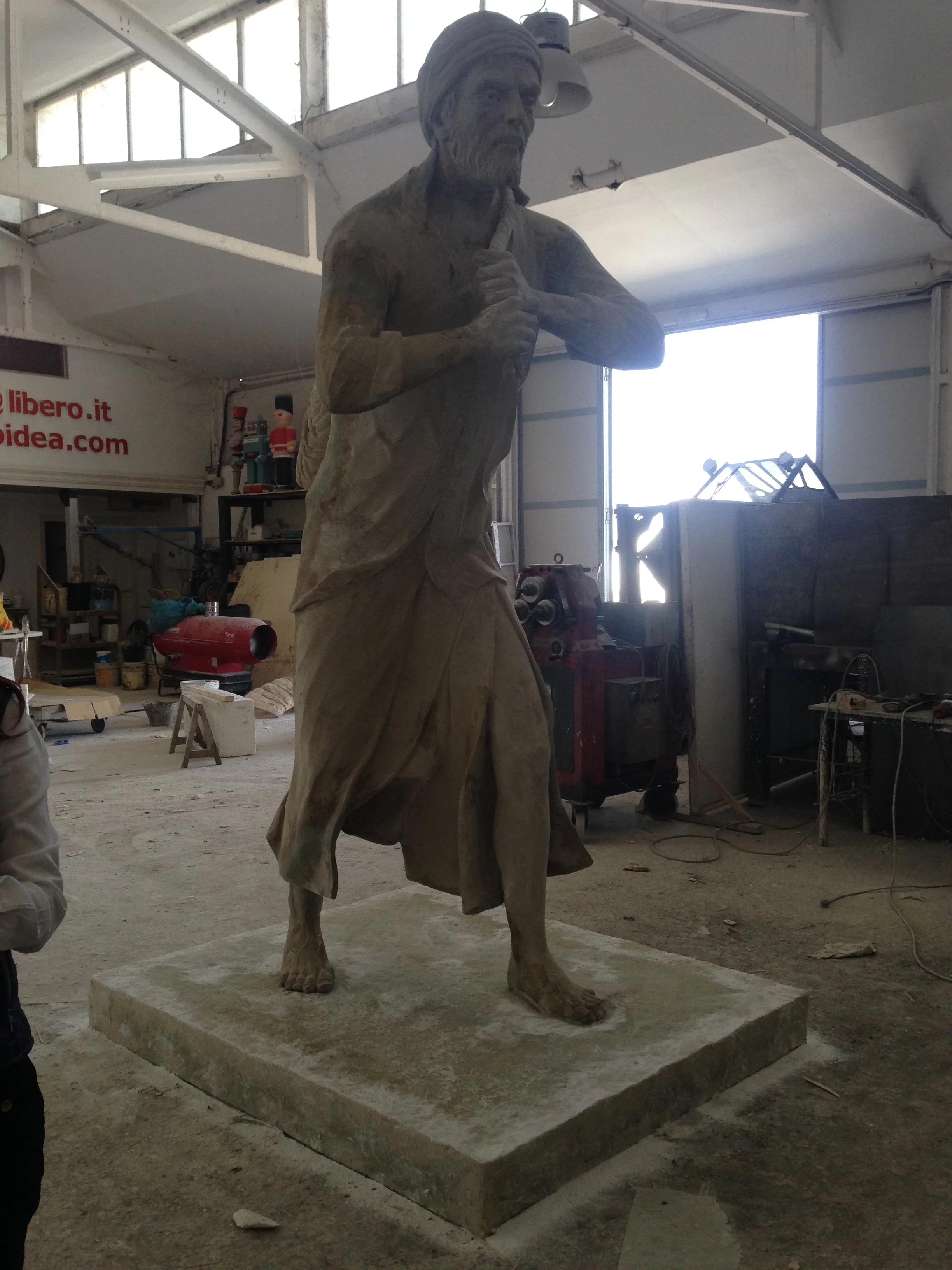 The statue of the Qatari man being sculpted in our workshops: life-size male figure in sand-coloured clay, fisherman's posture with one hand on the chest, draped garments sculpted in folds, keffiyeh modelled on the head; in the background the SCENO IDEA logo on the workshop wall and other sculptures in progress