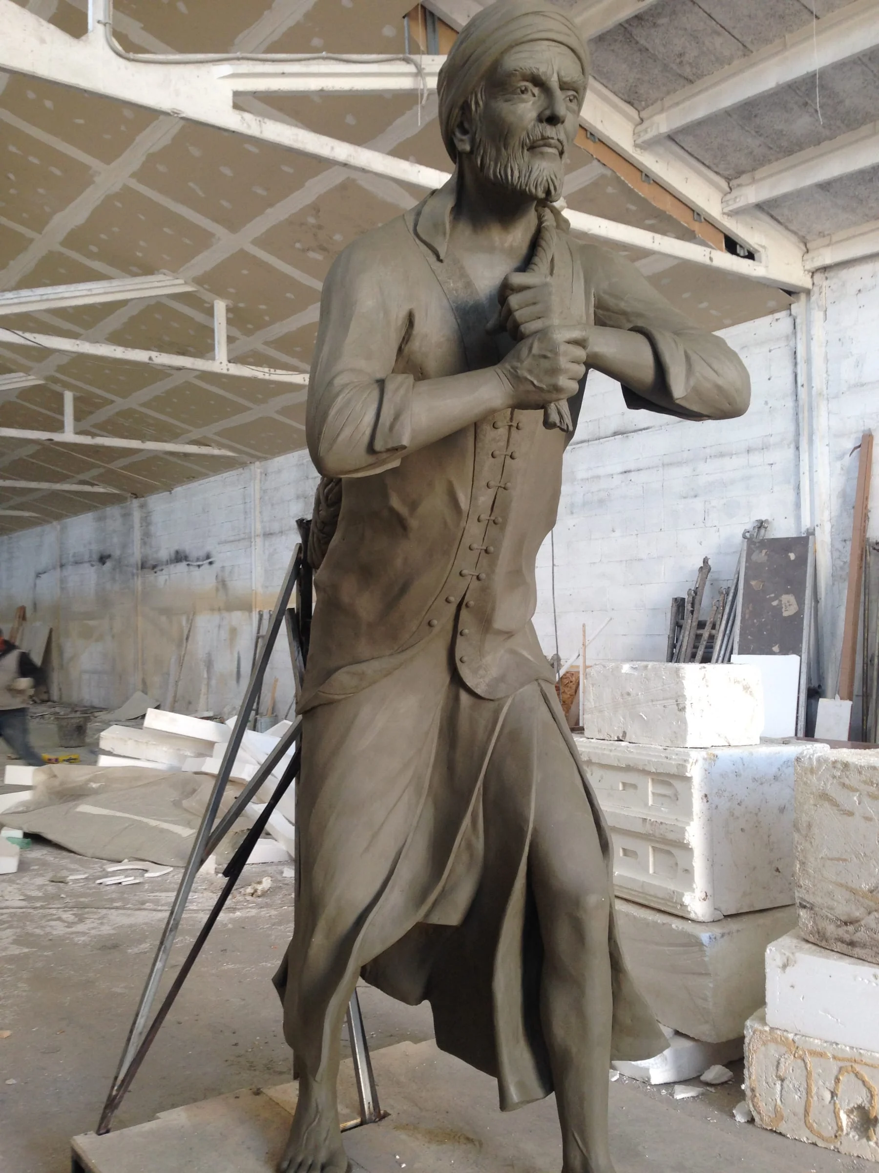 The same statue seen in profile: posture of the Qatari fisherman figure, drapes of the tunic modelled in folds, keffiyeh sculpted on the head, raw concrete base, workshop hall in the background