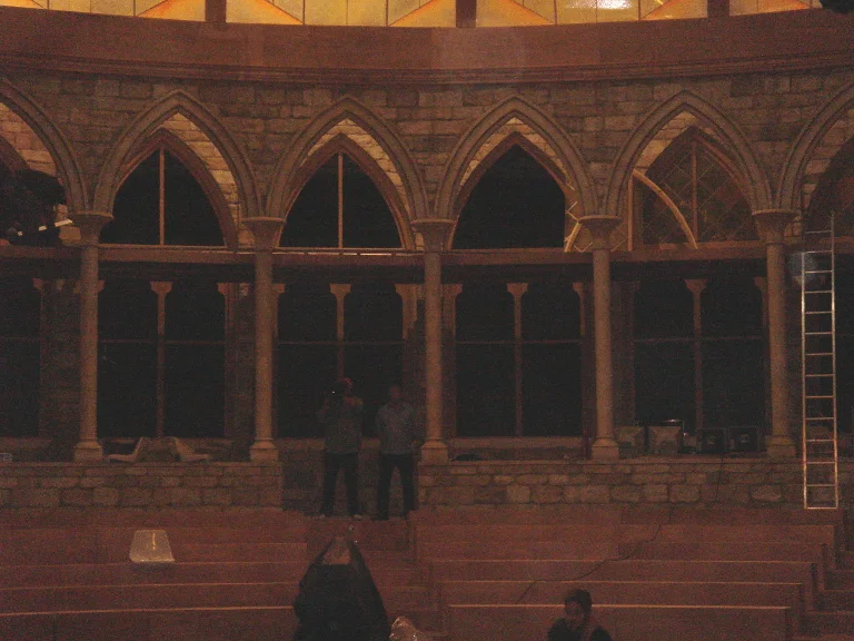 Detail of the gothic pointed arches of the Fattore C arena: sequence of arches on slender columns in grey faux stone with classical denticulation, a balustraded loggia above, the audience seating with red seats below, two people standing in the foreground for scale reference, night atmosphere of the final on-site stage