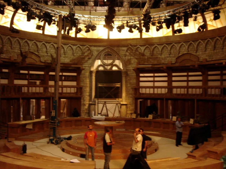 Panoramic view of the gothic circular arena of Fattore C in studio: upper dome with stage lighting, circular wall in double order — pointed arches in grey faux stone above, dark-wood lattice below, central arched entrance, light-wood circular stage at the centre with the final-stage crew, golden service lighting