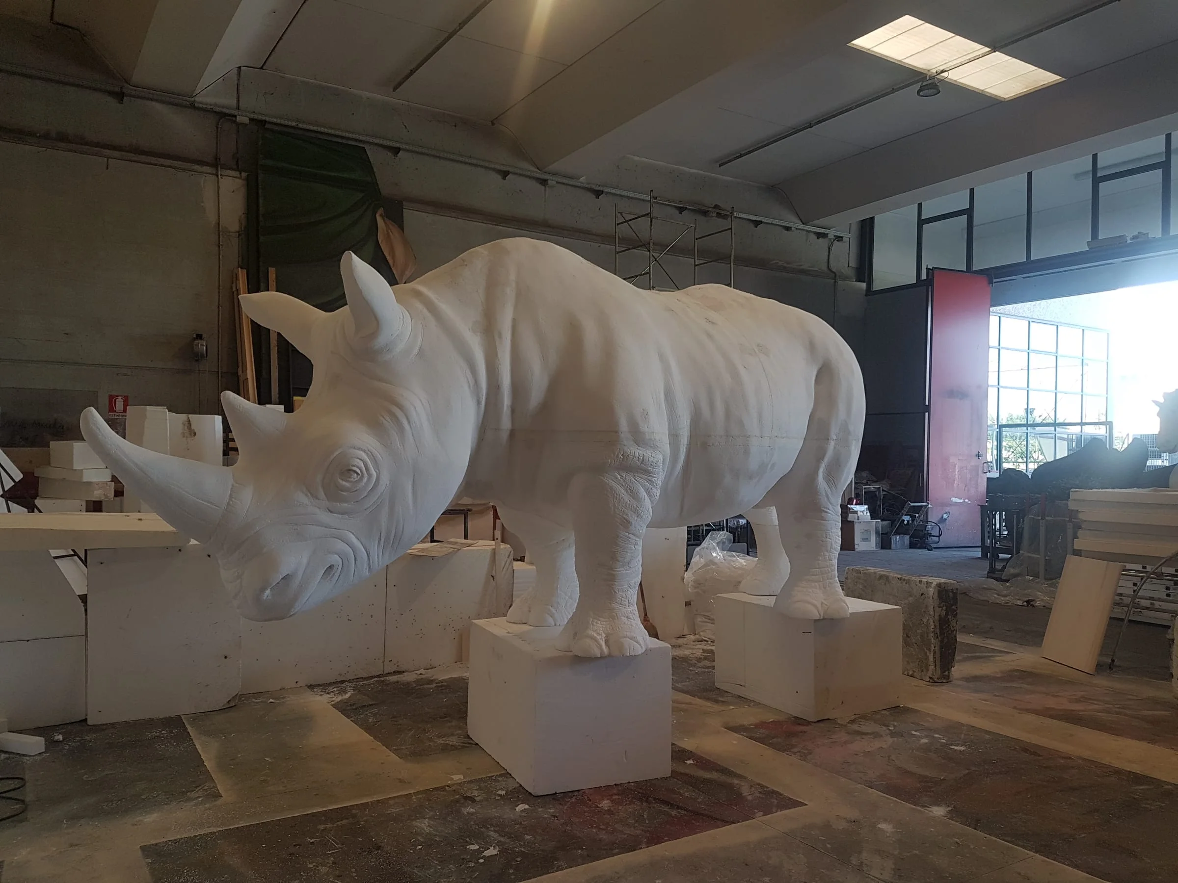 The life-size white rhinoceros sculpted by Creation Art for Fondazione Alda Fendi: frontal 3/4 view in the workshop, raw white polystyrene before the final finish, the four legs resting on polystyrene blocks, the folds of the skin and the eye sculpted by hand, industrial ceiling of the workshop, stained glass windows and polystyrene work blocks in the background