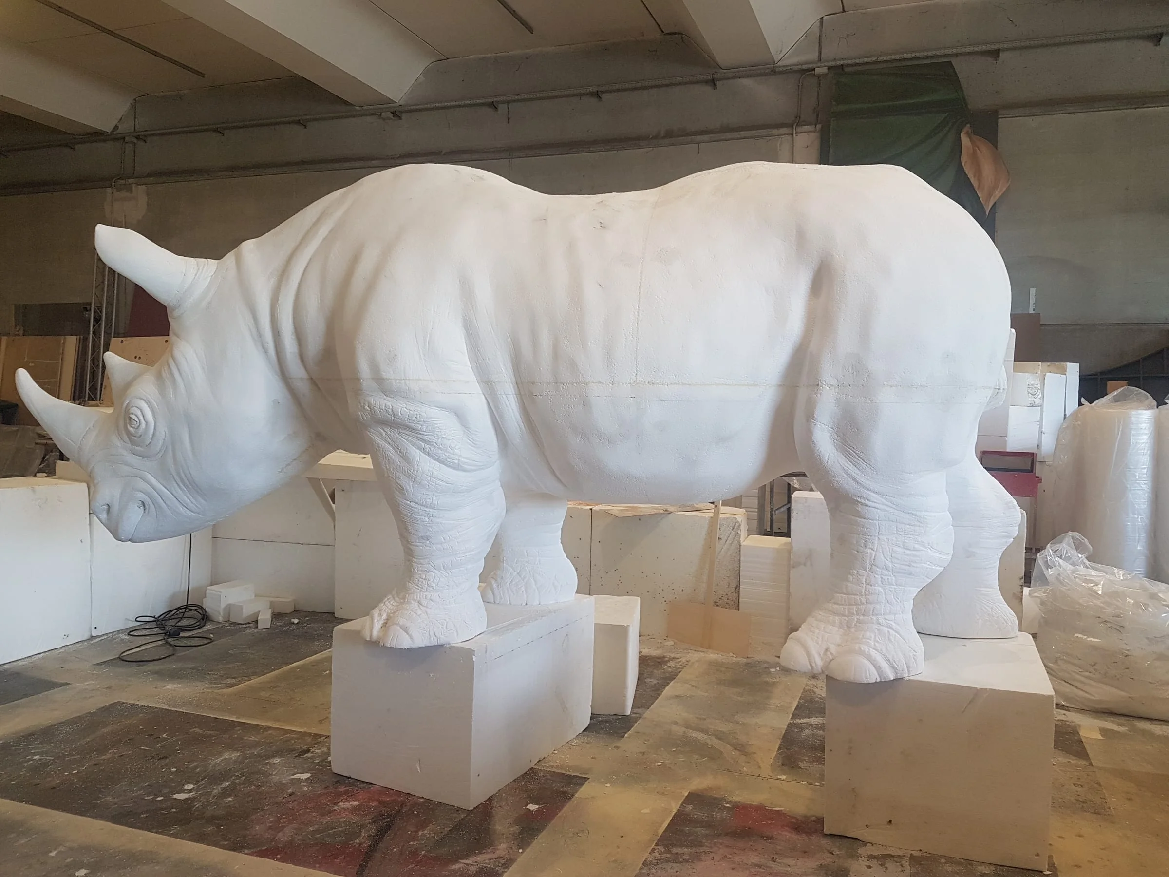 The same rhinoceros seen from behind/side: rear-side view, the curve of the back, the tail, the two sculpted horns, the massive legs on polystyrene blocks, the workshop floor with paint tiles, industrial ceiling of the Creation Art workshop