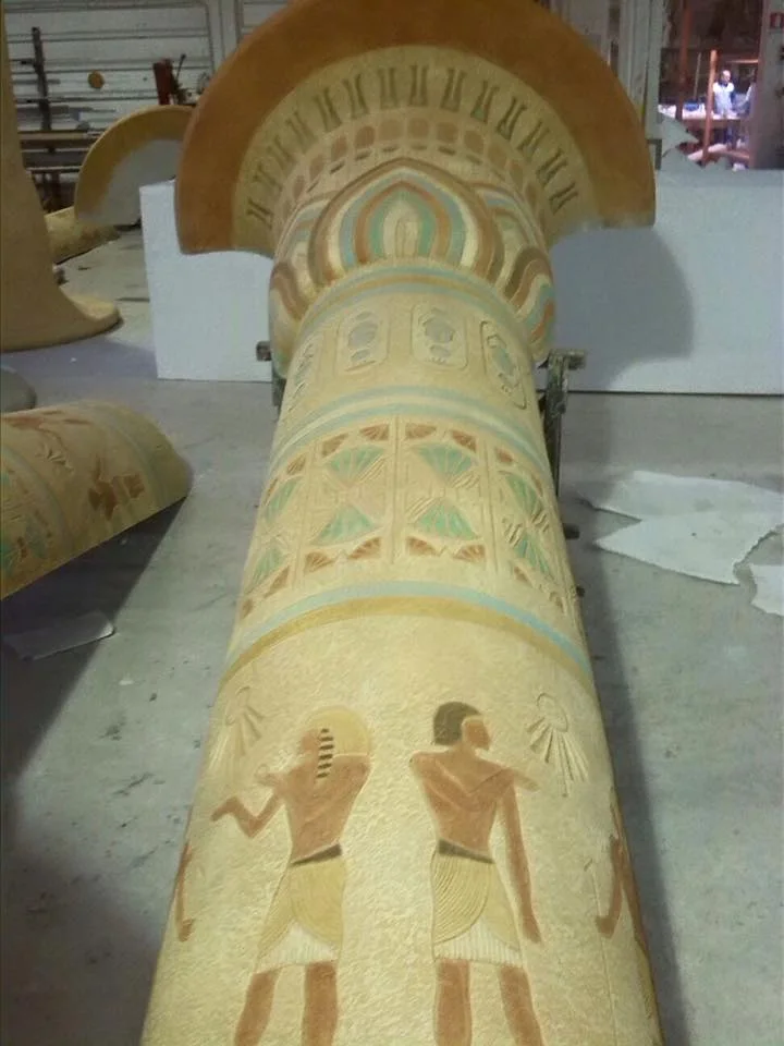Close-up detail of a finished papyriform column: papyrus-leaf capital painted with polychrome bands (gold, green, blue, pink), decorative bands with stylised hieroglyphs, at the bottom a cartouche with two figures of Egyptian offering-bearers sculpted and hand-painted