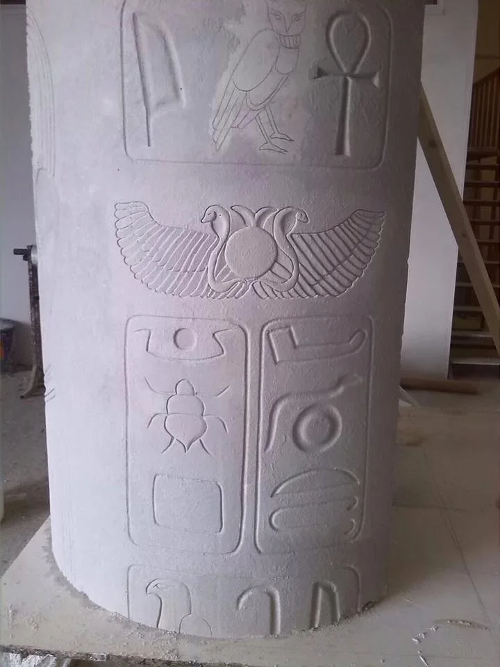 Egyptian column in freshly carved white polystyrene: at the top a royal cartouche with falcon-Horus and ankh, at the centre the winged sun decoration with side cobras, at the bottom another cartouche with scarab and hieroglyphic symbols; awaiting polychrome painting, with construction scaffolding in the background