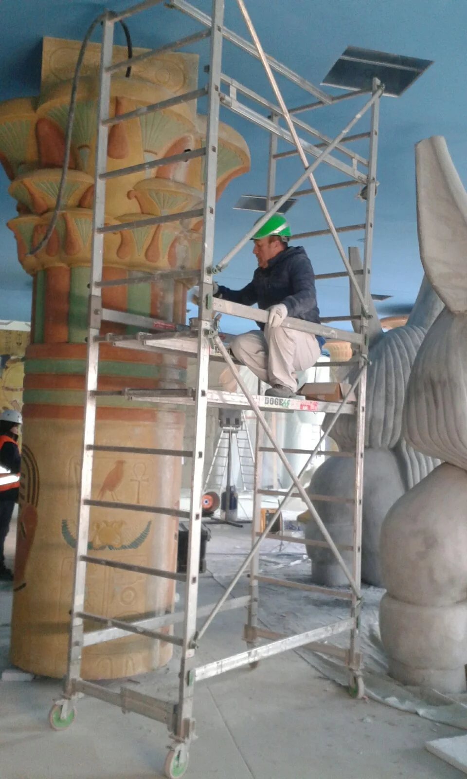 Construction of the Adventure Hotel lobby at Gardaland: a large papyriform column painted in orange, green and gold with hieroglyphic cartouches at the base and a papyrus-chalice capital on top; on the right a large grey-white Anubis statue in faux stone; a worker on the mobile scaffold is painting the capital, blue ceiling like an Egyptian sky