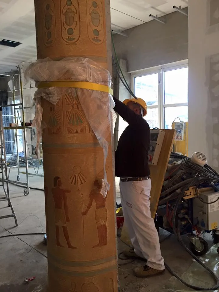 Creation Art worker with yellow helmet and white overalls painting an Egyptian column with coloured hieroglyphs: royal cartouches at the top with scarabs and cobras, central band with stylised papyrus decoration, below figures of Egyptian offering-bearers; in the background another column wrapped in protective plastic, hotel windows