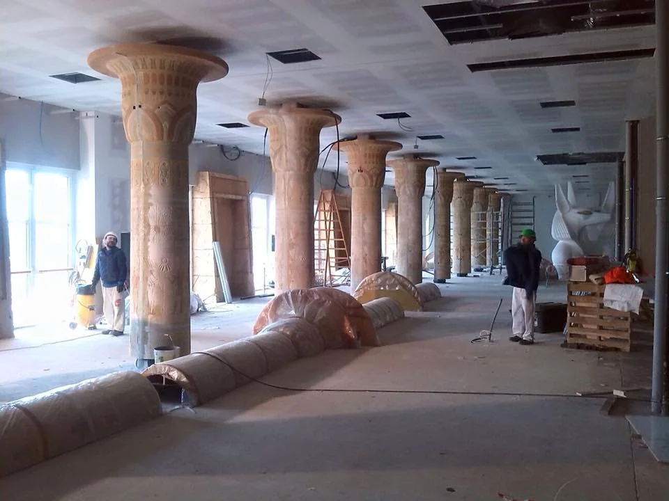 Construction of an Adventure Hotel hall: six tall Egyptian papyriform columns painted in polychrome stripes (pink, orange, gold, blue), papyrus-leaf capitals, two workers with helmets at work between the columns, a large white Anubis statue in the background on the right being installed, the construction floor with pipes and conduits laid out