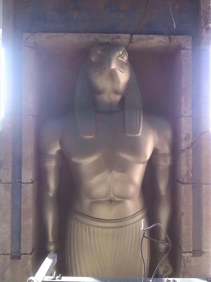 Horus statue (falcon-headed god with human body) finished in bronze colour in the stone niche of the Adventure Hotel: arms crossed on the chest in solemn pose, sculpted pleated Egyptian robes, above the niche a frieze decorated with golden hieroglyphs on black background