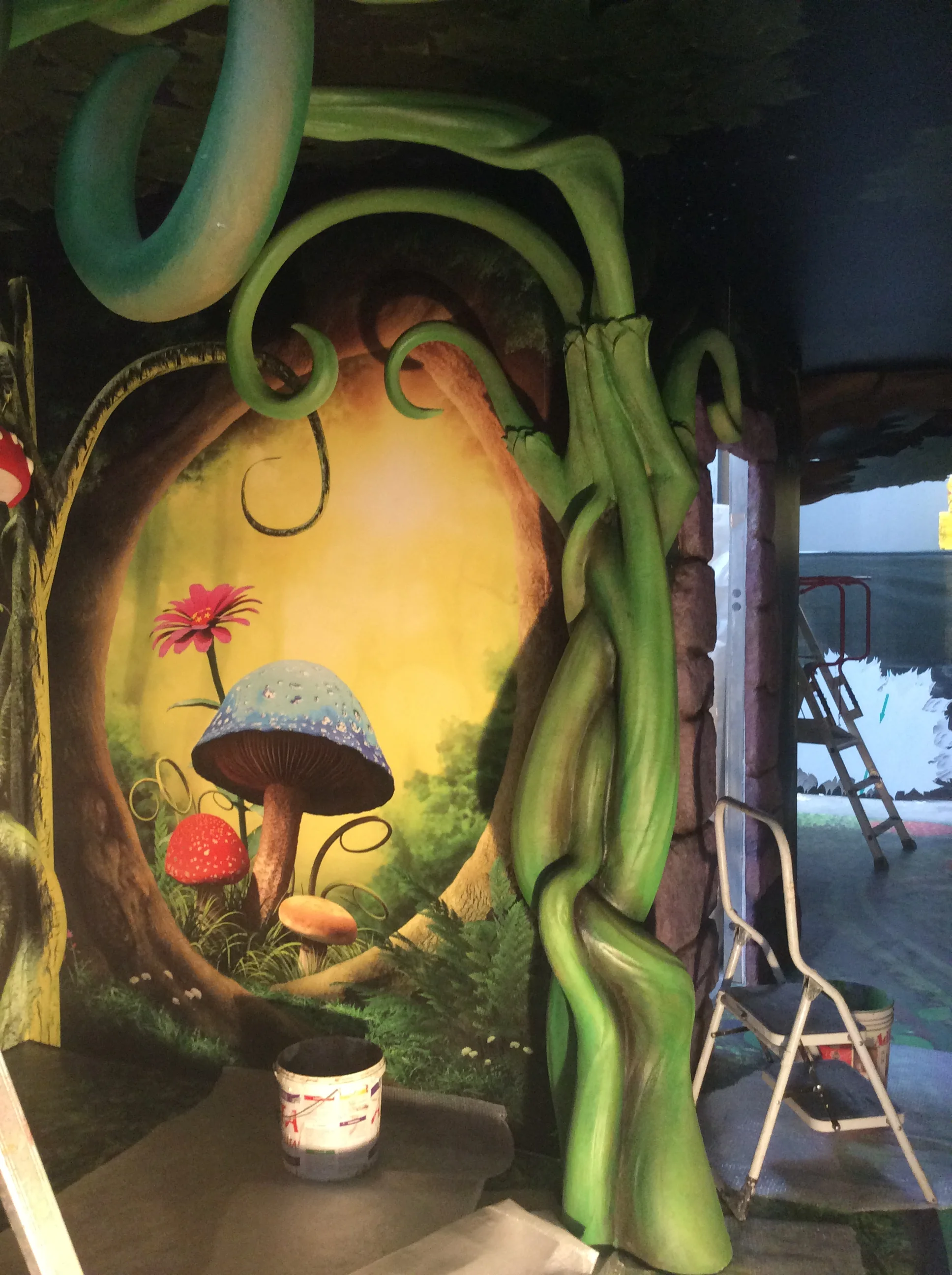 Forest room of the Magic Hotel: porthole painted on the wall opening onto a cartoon forest scene with blue-red polka-dot mushroom, red strawberry, pink flower, yellow sun in the background; around the porthole giant green tendrils sculpted in full round descend from the ceiling framing the scene