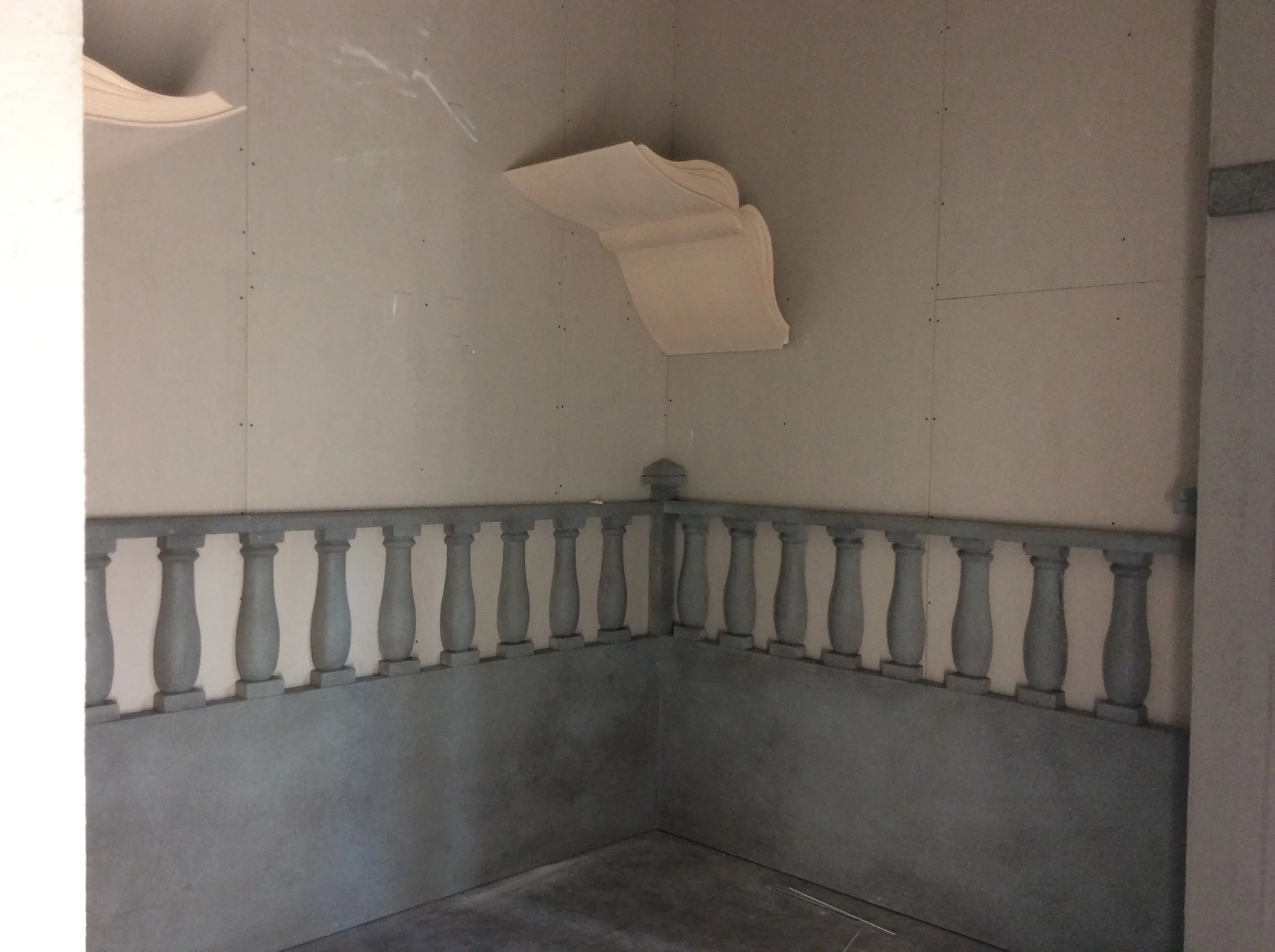 Wizard room of the Magic Hotel under construction: balustrade in grey faux stone sculpted with turned spindles along the base of the walls, scrolls sculpted hanging from above like medieval trophies, drywall walls still to be plastered