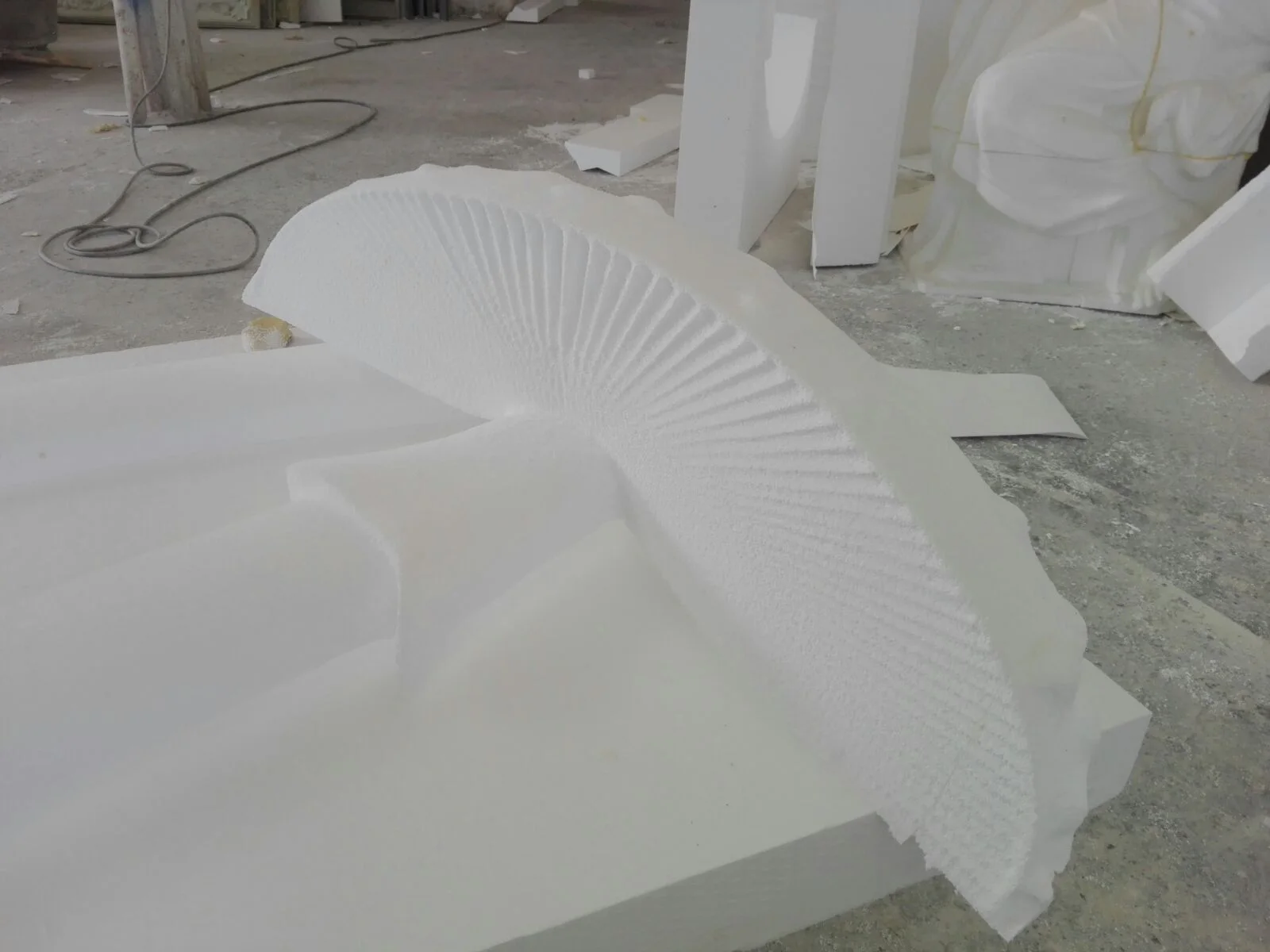Large fan-shaped shell sculpted in raw white polystyrene: semicircular shape with deep radial striations from the centre, placed on the workshop floor awaiting finish