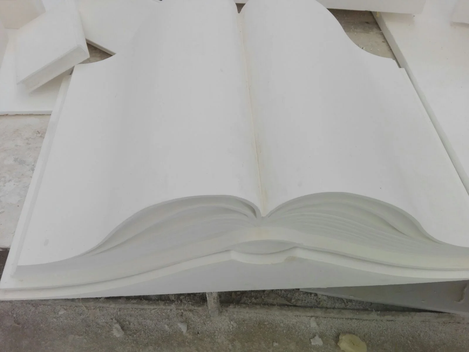 Giant open magic book sculpted in raw white polystyrene: pages half-flipped with the spine at the centre, page-edge section visible from the side like an old book, dimensions about two metres wide; being prepared in our workshops before painting