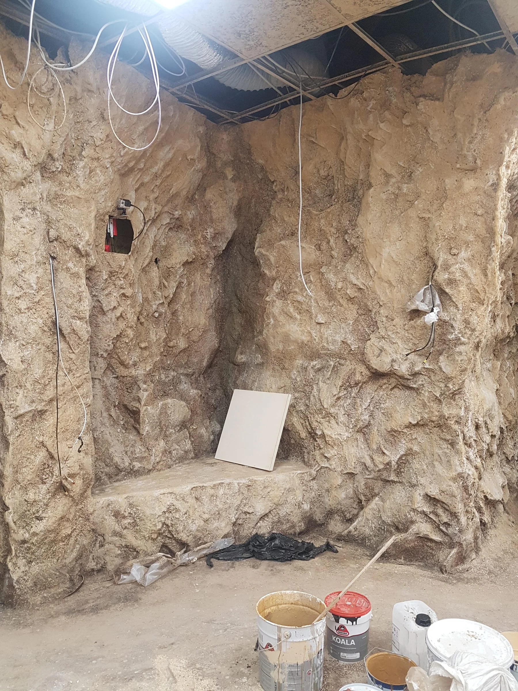 Workshop: rock walls still in progress, sandy ground, buckets of coloured mortar on the right, electrical cables on the ground. Rough texture of the fake stone still to be patinated