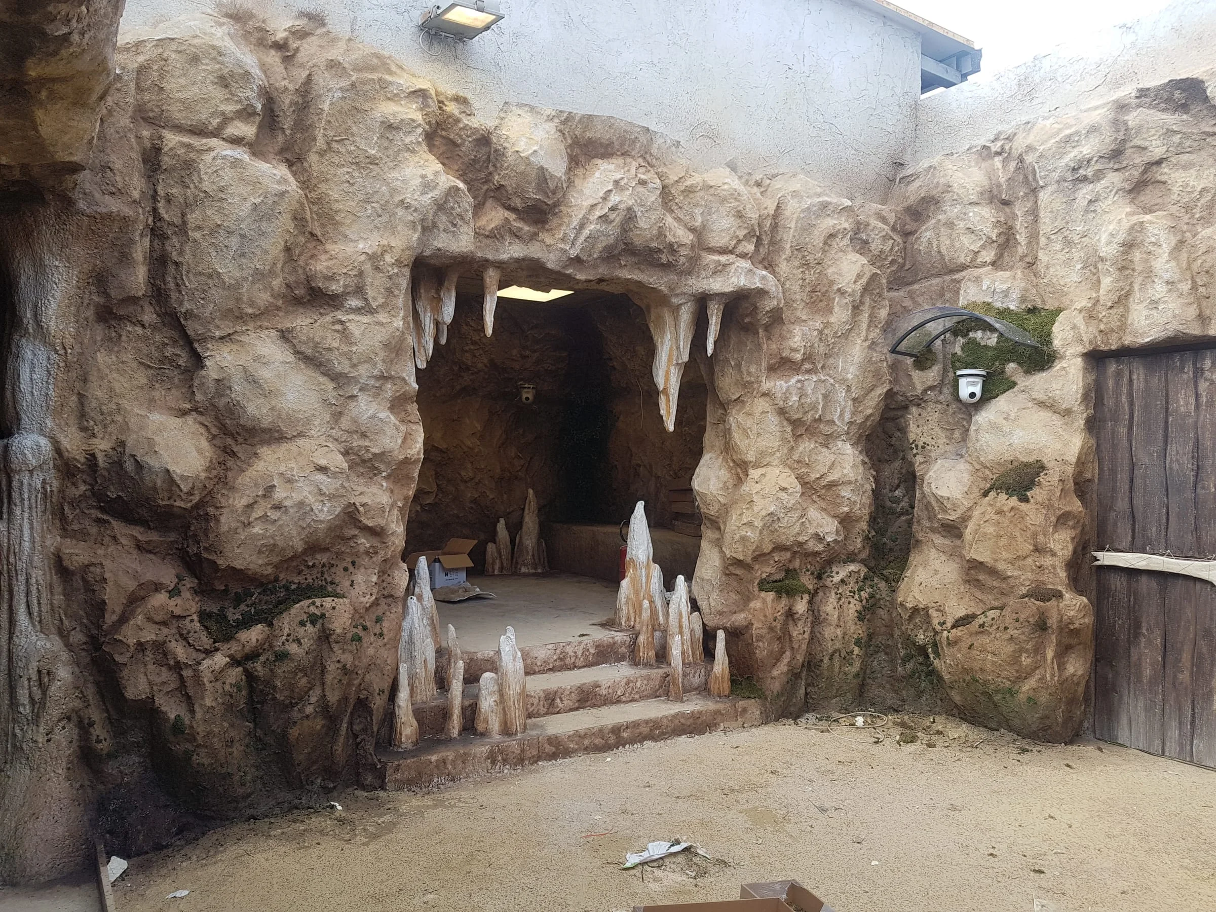 Front view of the Cave: old wooden double door between sculpted rock walls, short stalactites hanging from the lintel, fake vegetation on the sides, sandy floor. Light streams in from the roof of the Cinecittà sound stage
