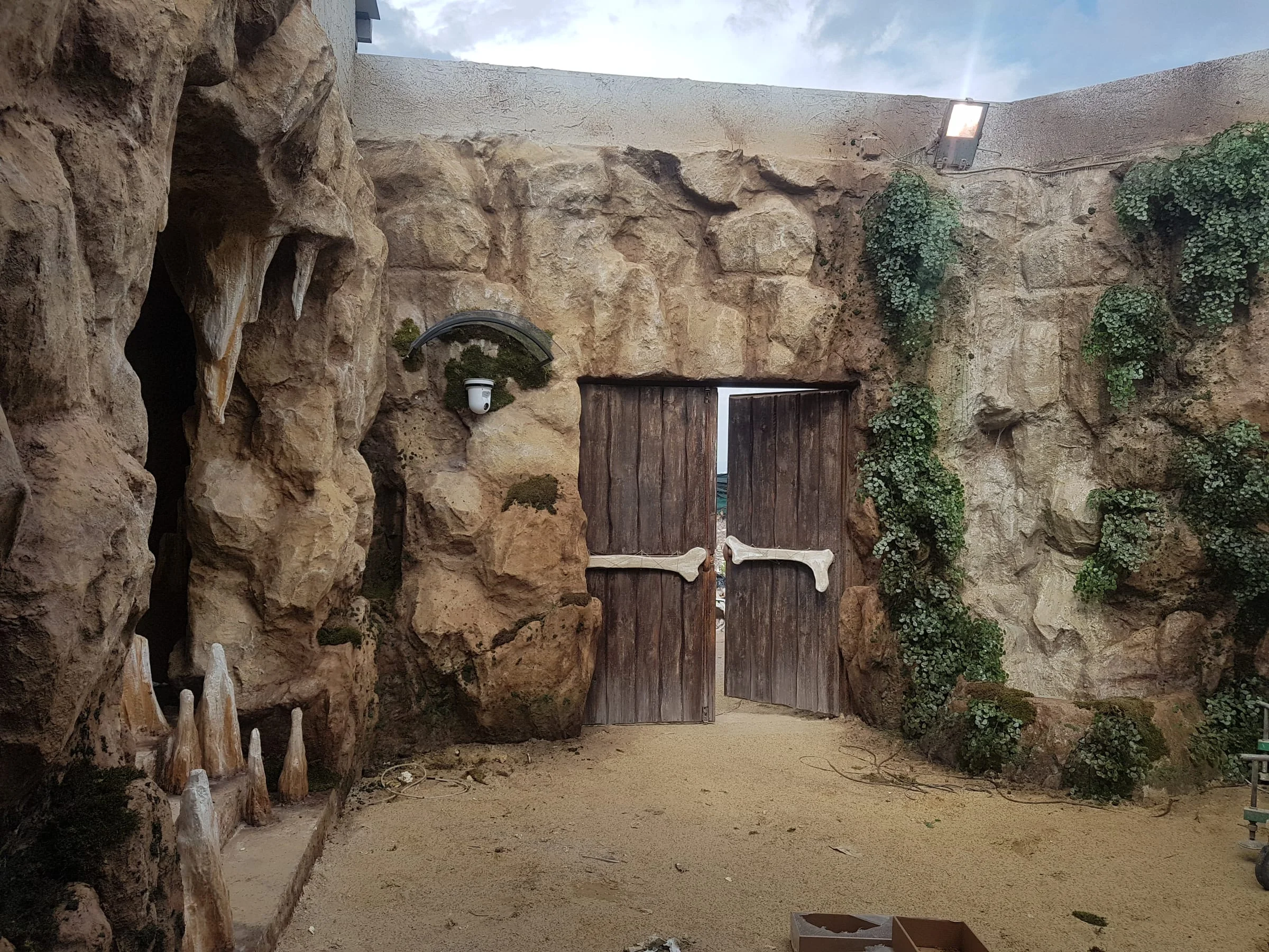 Three-quarter view of the Cave with sculpted stone steps rising toward an arched opening, lateral stalactites and stalagmites, a fake street lamp resting at the top, fake hanging vegetation