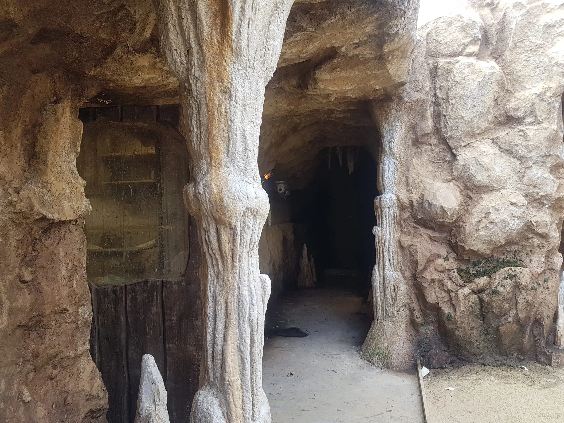 Interno della Caverna del set GF VIP 3 a Cinecittà: stalattiti pendenti dall'alto del teatro di posa, doppia porta antica in legno, vegetazione finta lungo le pareti rocciose scolpite